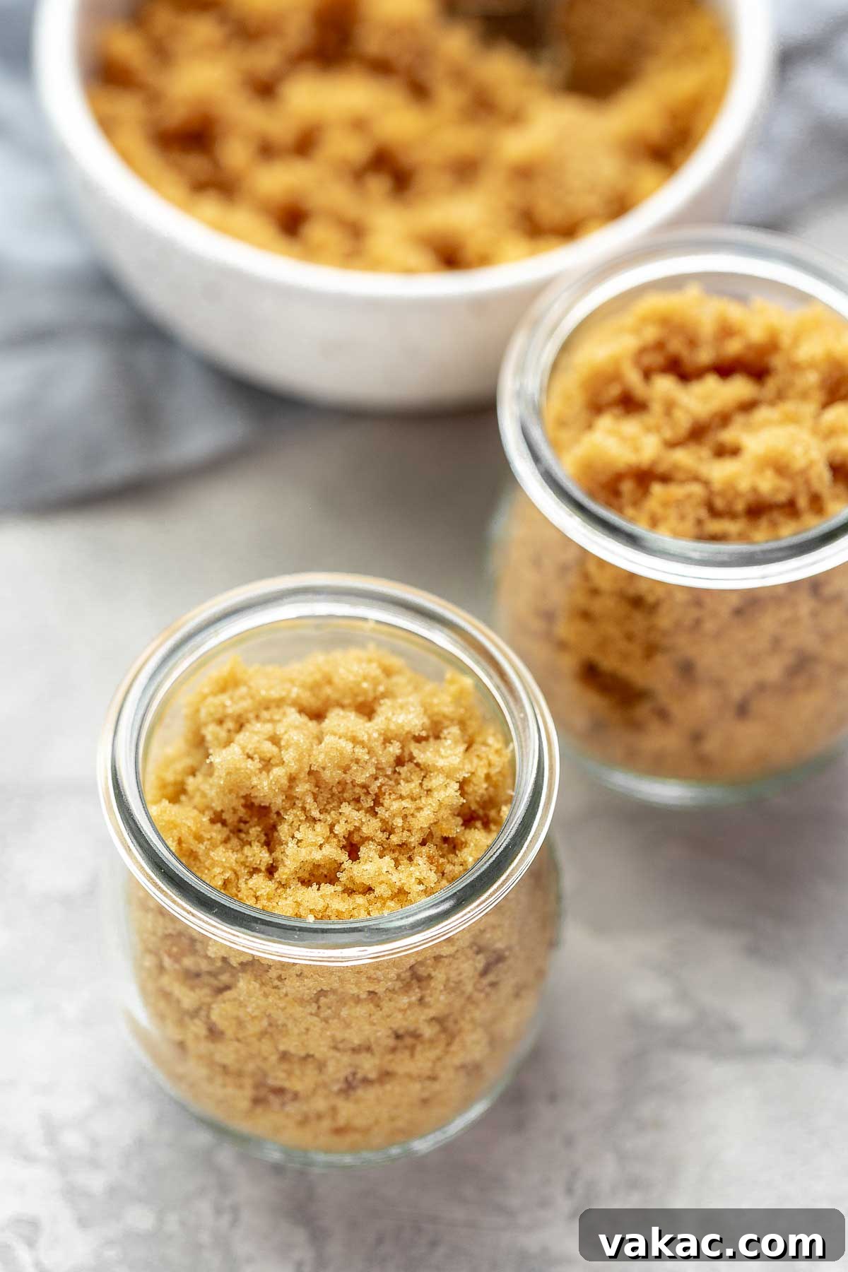 Two glass jars filled with homemade brown sugar substitute. A bowl of brown sugar is in the background, suggesting a comparison.