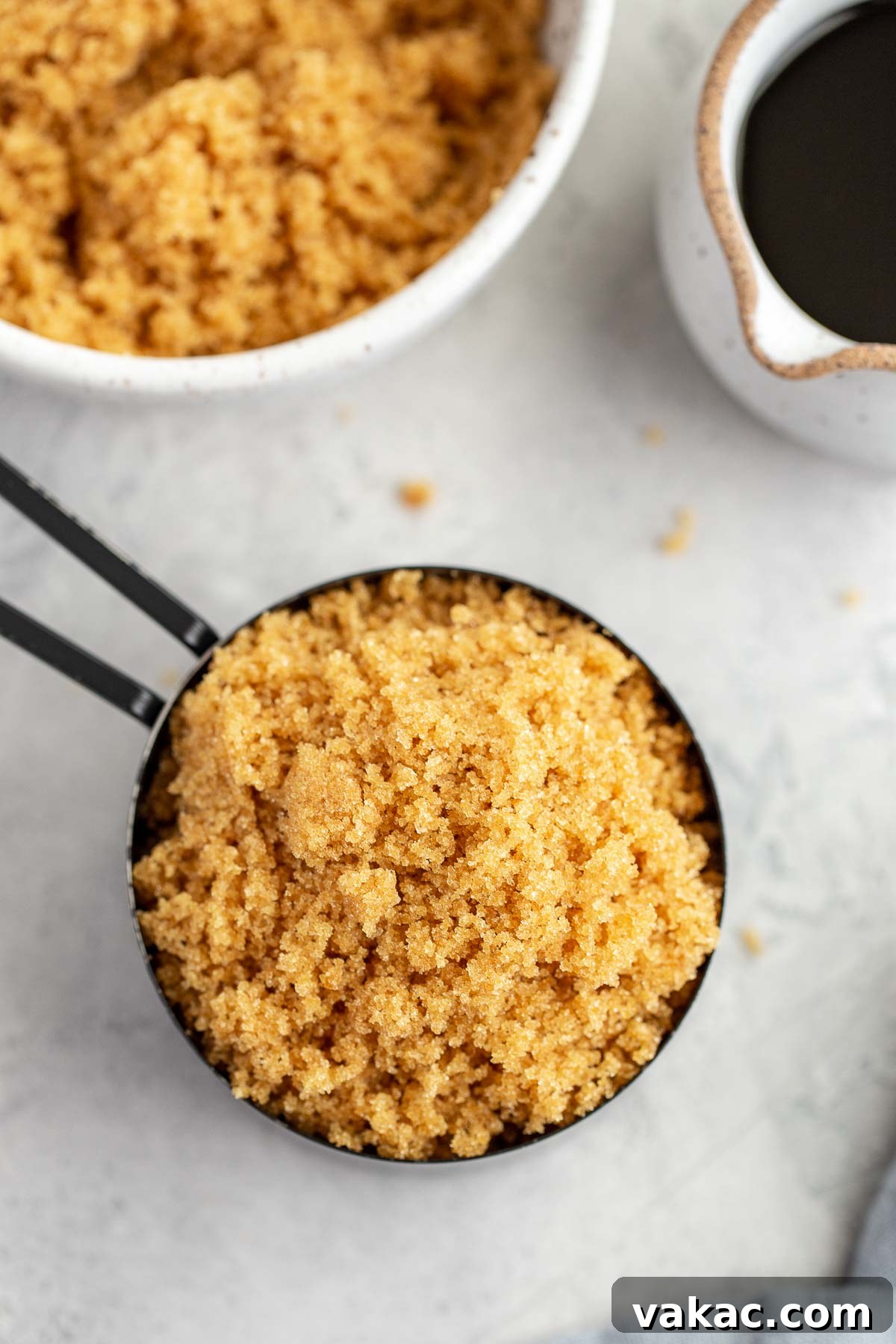 An overhead view of a measuring cup filled with homemade brown sugar, with a bowl of brown sugar and a small jug of molasses nearby.