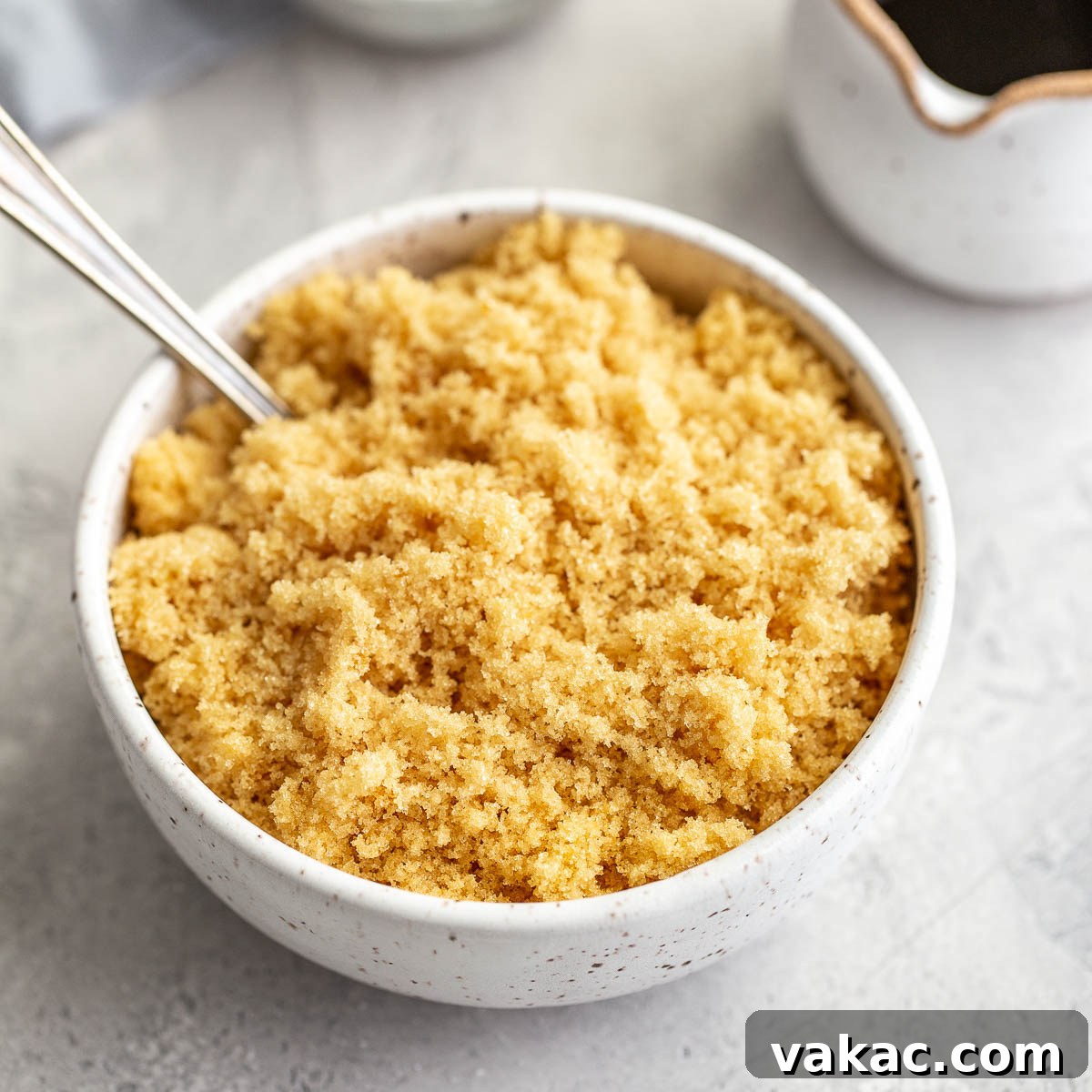 A bowl of light brown sugar with a spoon resting in it, showcasing the texture. A small container of molasses sits in the background.