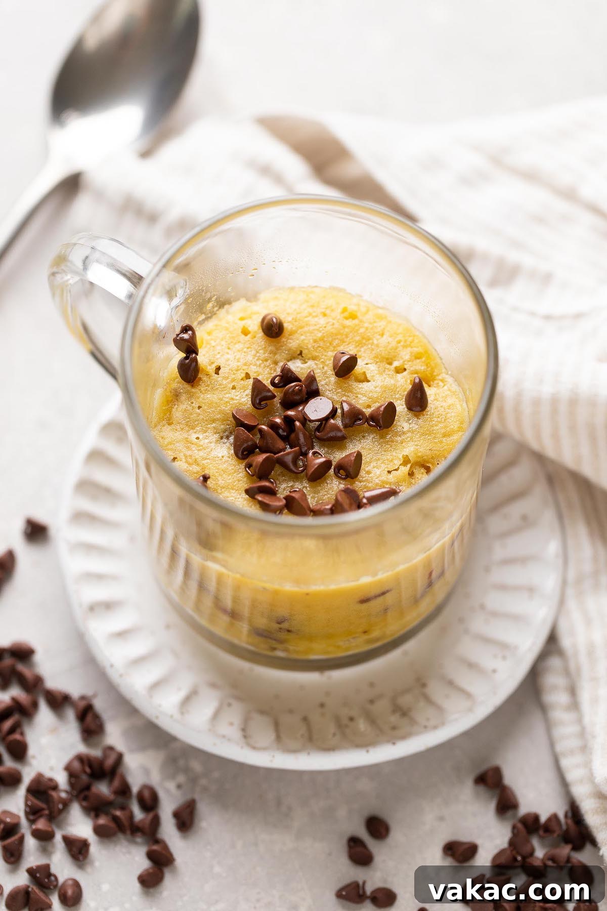 Chocolate Chip Mug Treat 2 A warm, gooey chocolate chip cookie in a mug, nestled on a white dessert plate. Miniature chocolate chips are sprinkled on top, with a spoon resting invitingly in the background, ready to dive in.