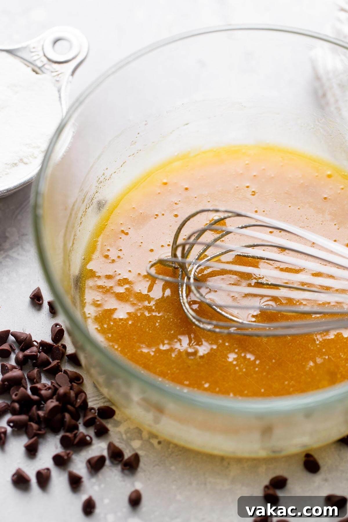 A close-up of melted butter and both granulated and brown sugars being whisked together in a clear glass mixing bowl, capturing the initial step of combining wet ingredients.