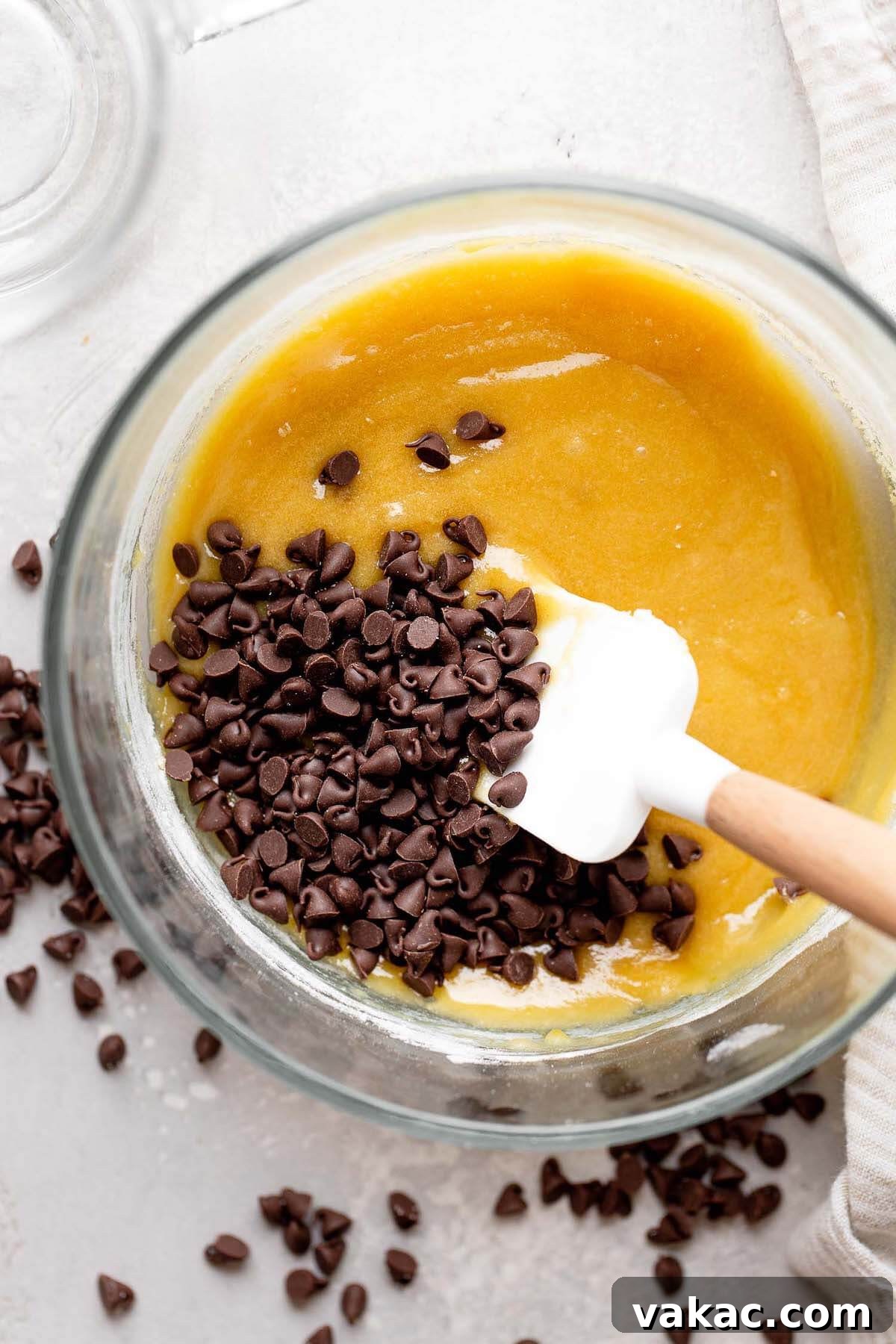 An overhead view of mini chocolate chips being gently folded into raw, rich cookie dough with a rubber spatula in a glass bowl, showcasing the final mixing step.