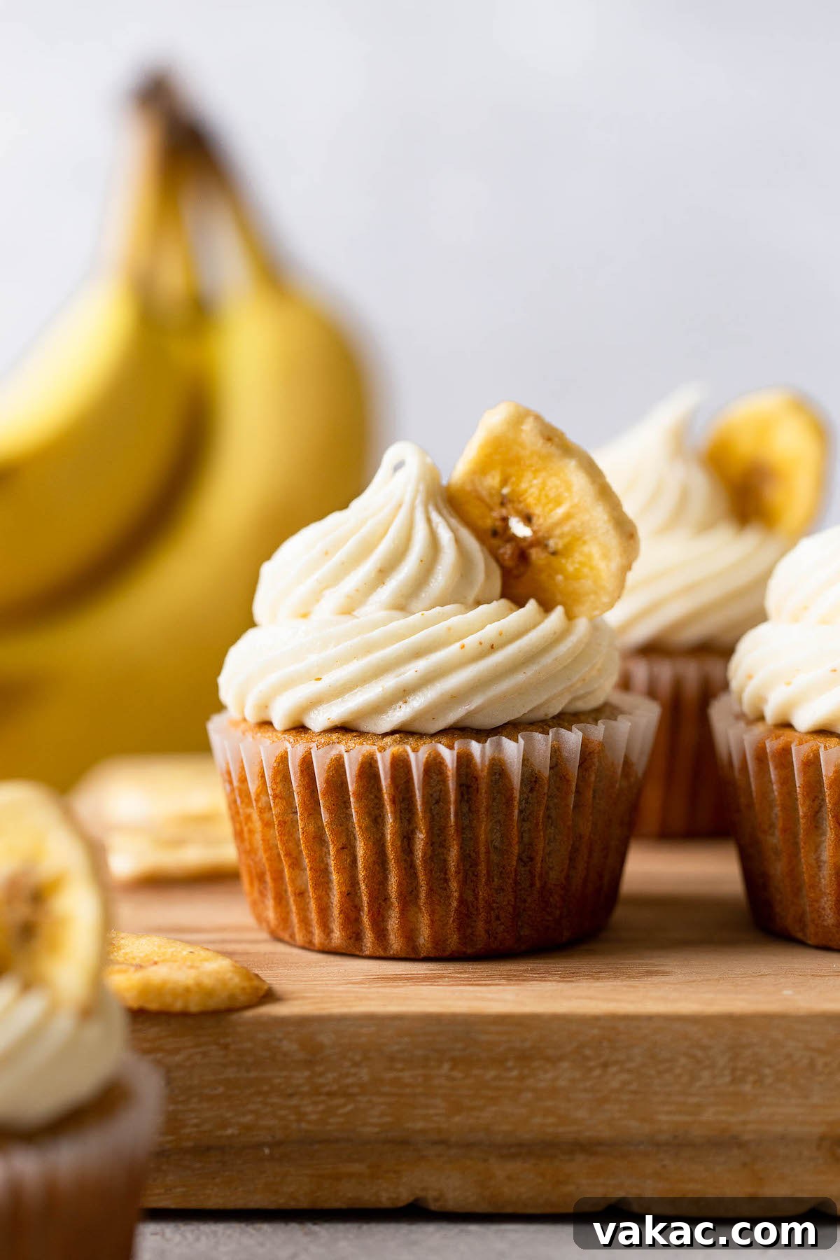 Sweet Banana Bites 2 Side view of banana cupcakes with cream cheese frosting on a wooden board. A bunch of bananas rest in the background.
