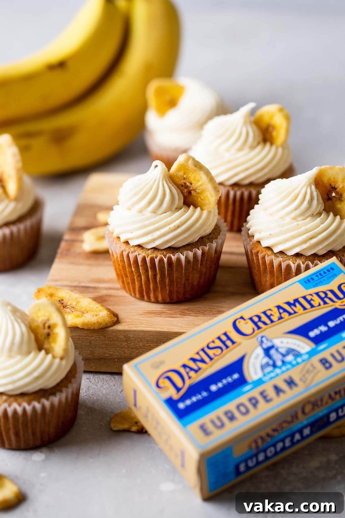 Sweet Banana Bites 6 Side view of banana cupcakes with cream cheese frosting on a wooden board. A bunch of bananas rest in the background.