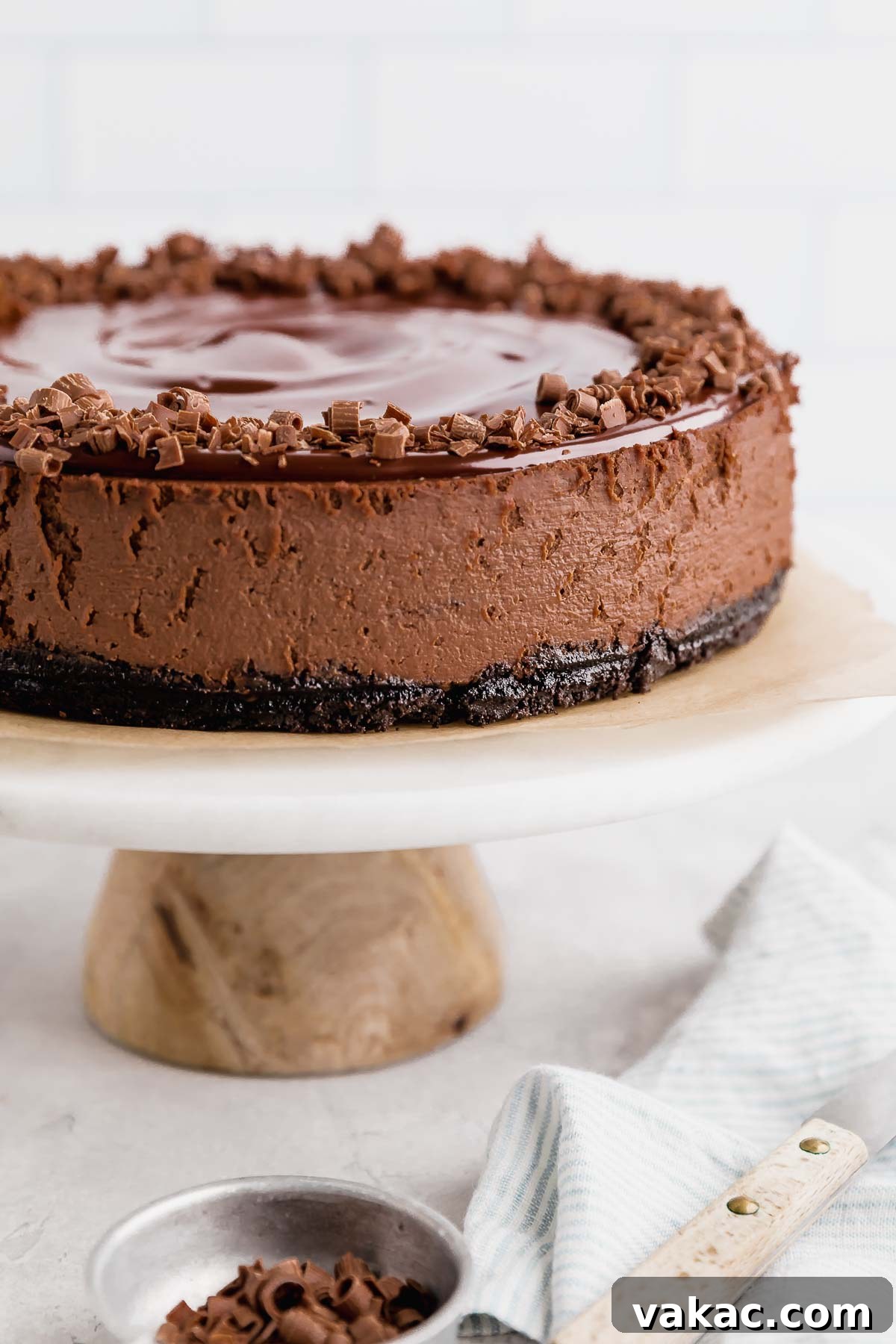 A side view of a chocolate cheesecake on a white marble cake stand. A small bowl of chocolate shavings rests in the foreground. The cheesecake is topped with chocolate ganache and chocolate curls, looking incredibly rich and inviting.