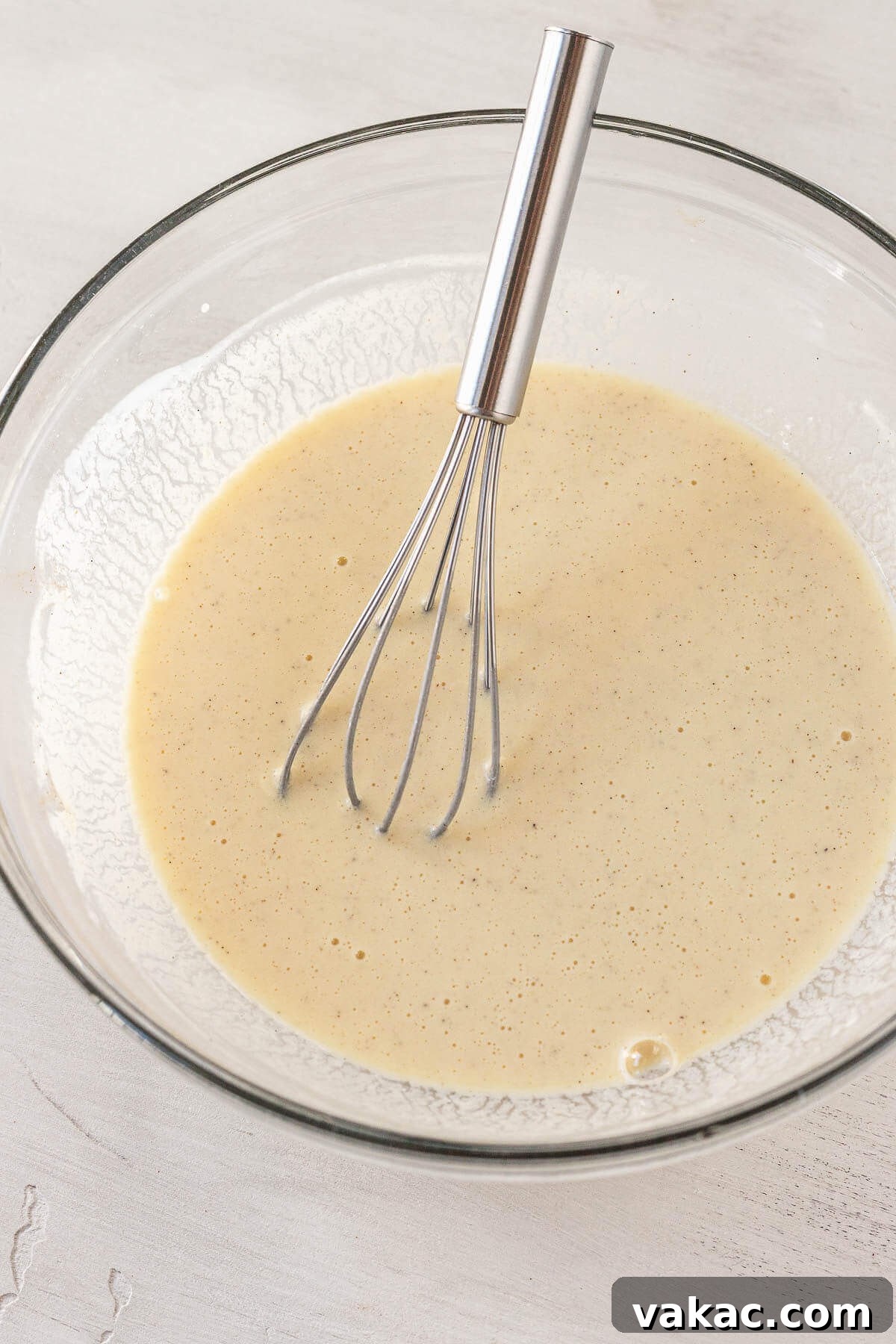 Buttermilk, sour cream, eggs, and vanilla mixed together in a clear glass mixing bowl with a metal whisk, showcasing the creamy wet ingredients for the cake batter.