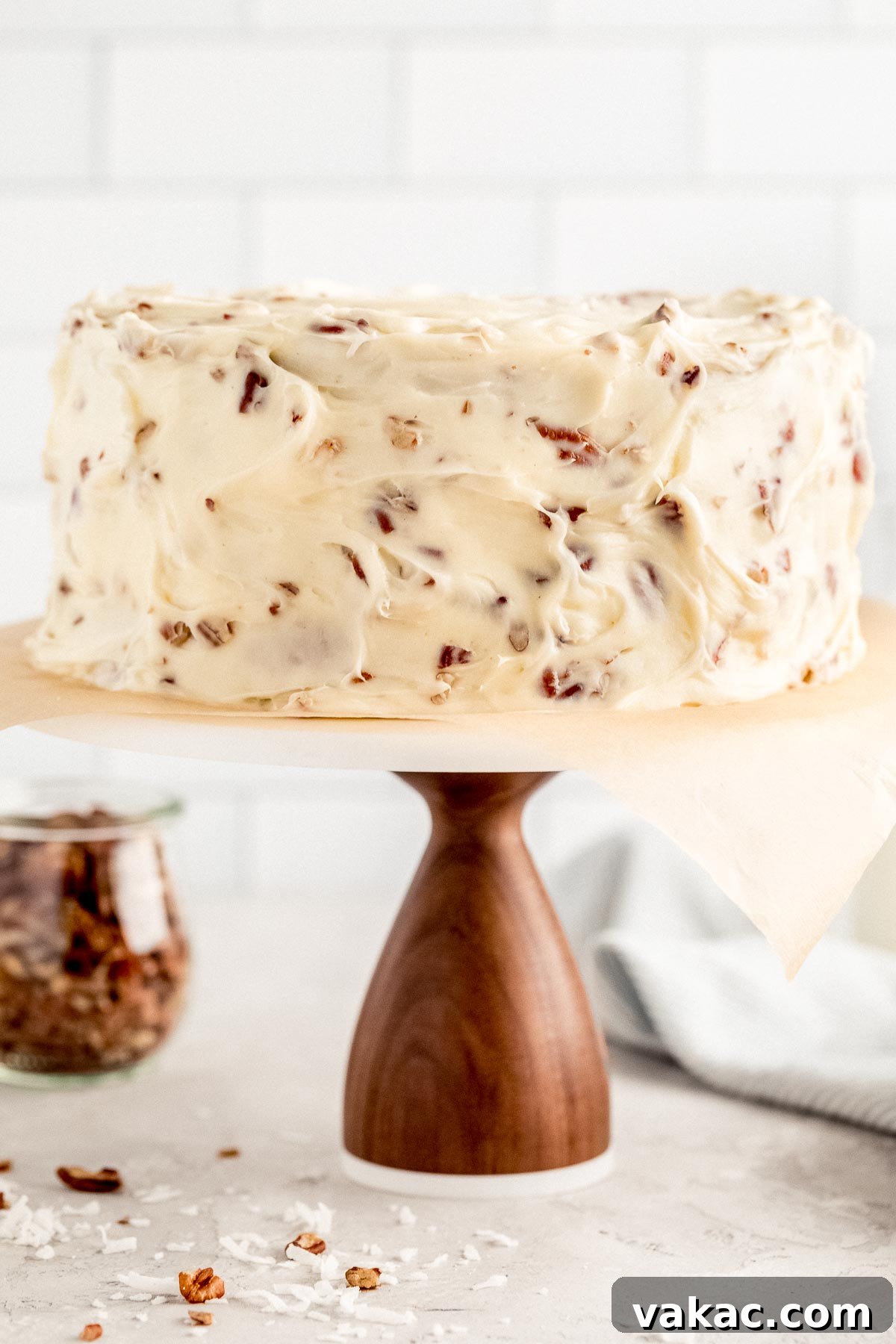 Velvet Italian Cream Indulgence 2 A stunning Italian Cream Cake adorned with a thick layer of cream cheese frosting and chopped pecans, displayed elegantly on a wooden cake stand with a marble top. A jar of extra chopped pecans and two vintage milk jugs are visible in the soft-focus background, enhancing the rustic, home-baked feel.
