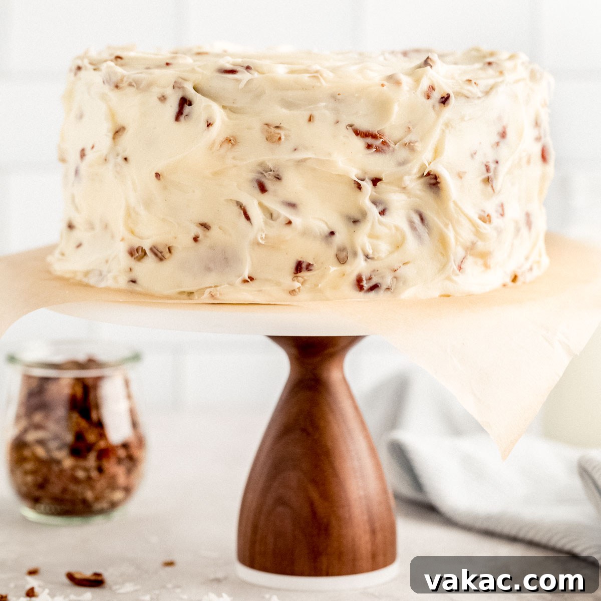 A rich, multi-layered Italian Cream Cake on a wooden stand with a marble top. The cake is generously frosted with cream cheese frosting and sprinkled with pecans. In the background, a jar of chopped pecans and milk jugs complete the cozy scene.
