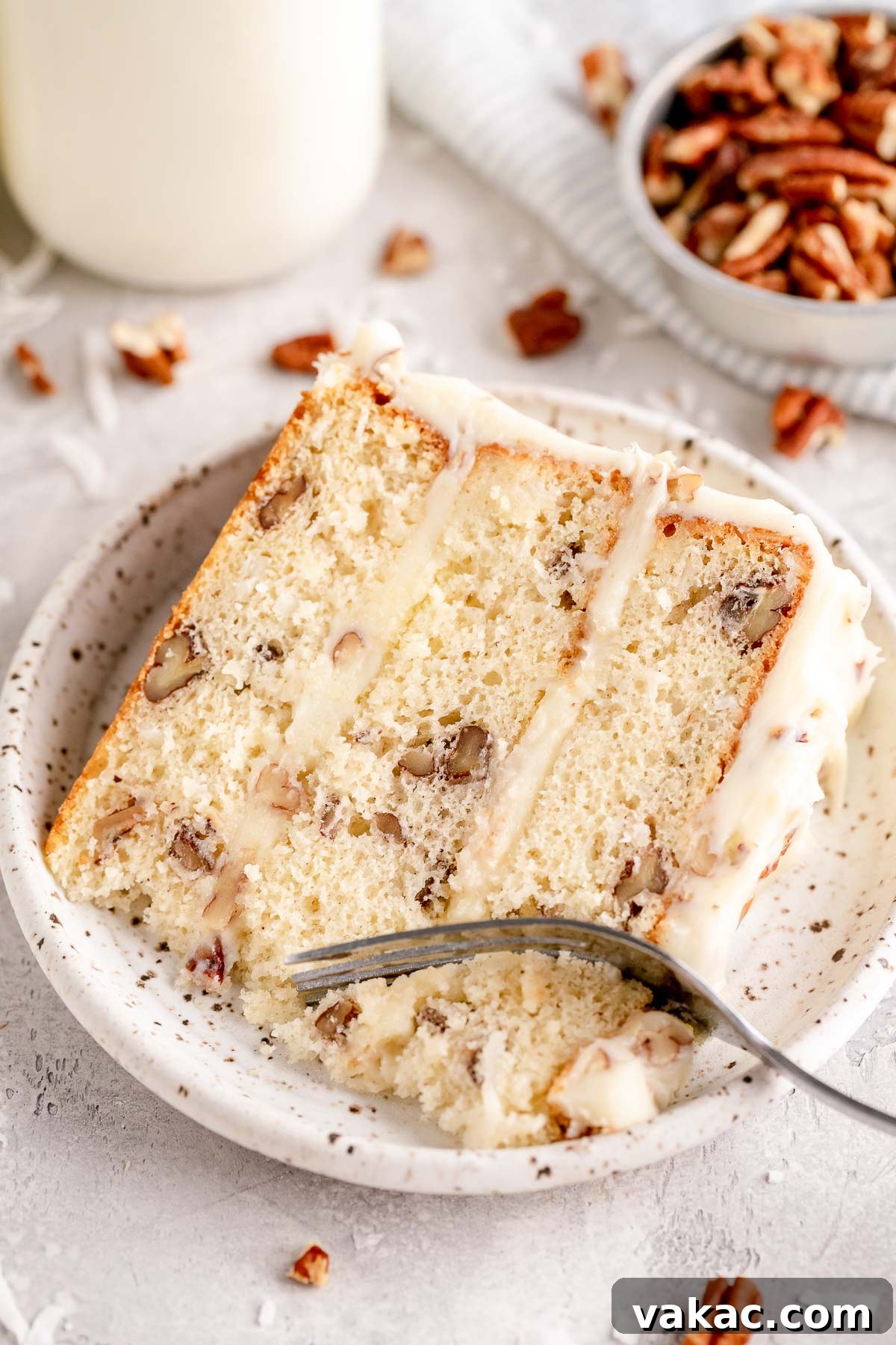 Velvet Italian Cream Indulgence 10 A delectable slice of Italian cream cake resting on a white speckled plate, showcasing its moist layers and generous cream cheese frosting. Scattered around the plate are fresh pecans and shredded coconut, highlighting the key ingredients of this rich dessert.