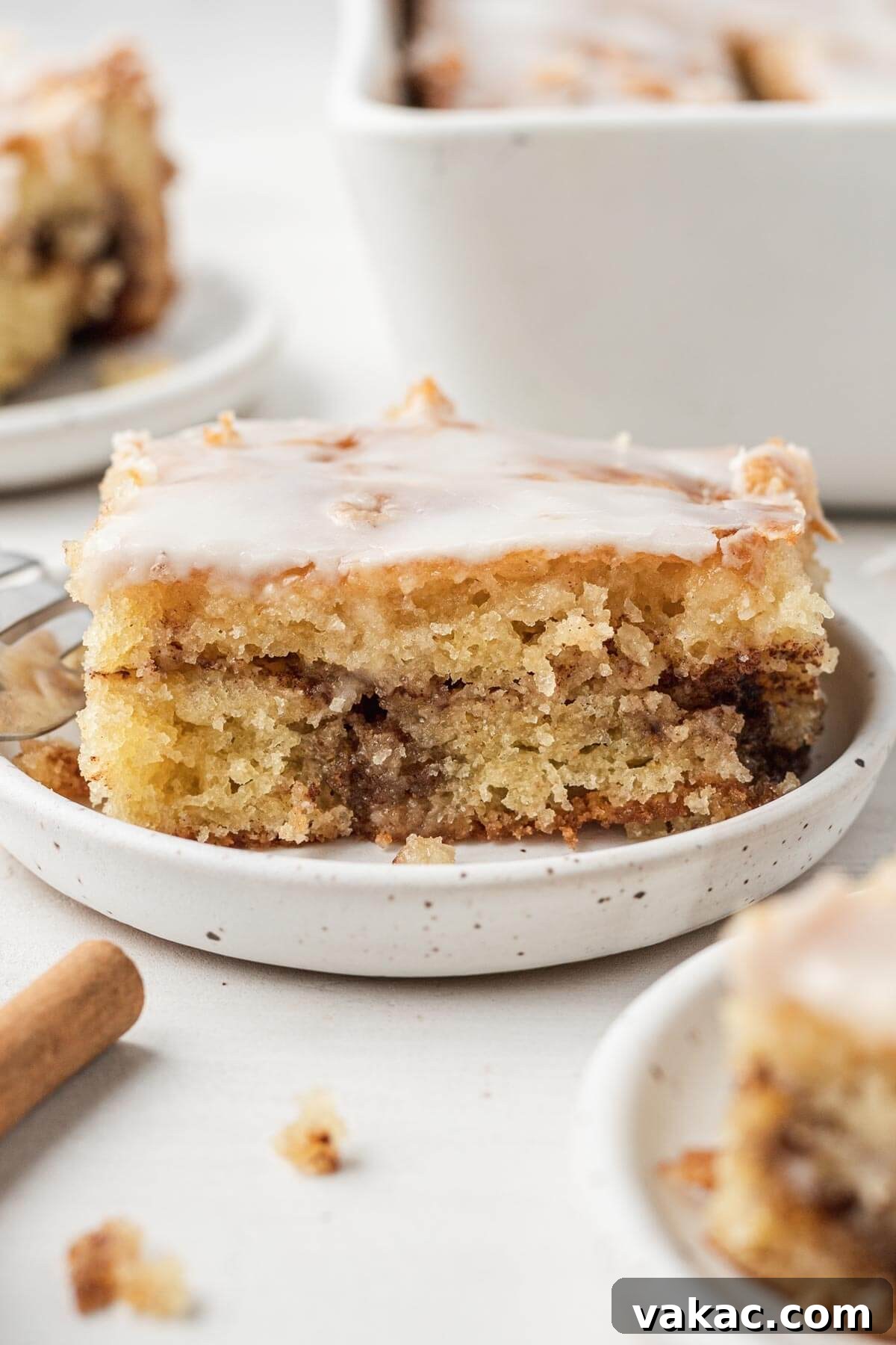 Cinnamon Swirl Honey Bun Delight 2 A slice of iced honey bun cake on a speckled white dessert dish. Additional cake slices rest in the foreground and background.