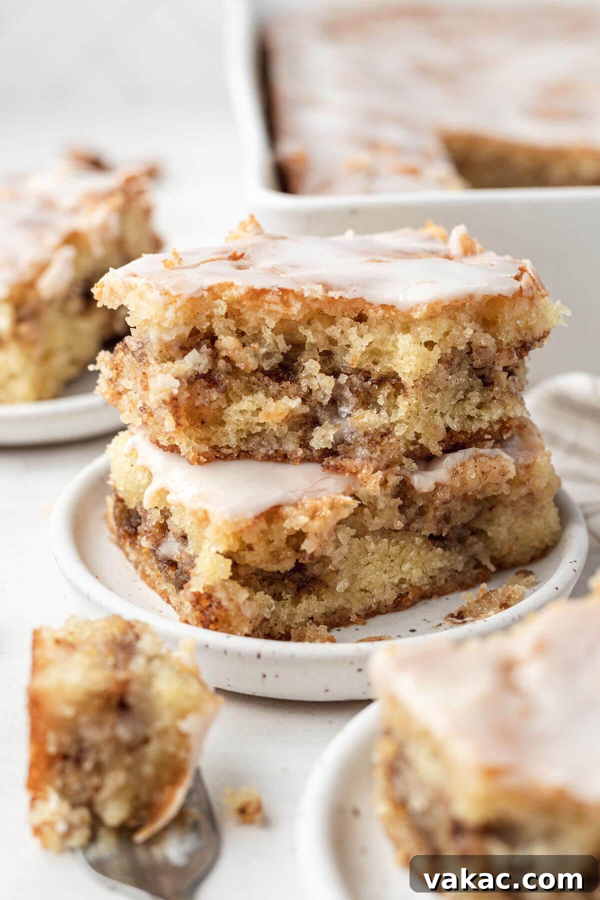 Cinnamon Swirl Honey Bun Delight 8 Two slices of honey bun cake, stacked on a white speckled dessert plate. Additional cake slices and a baking pan rest in the foreground and background.