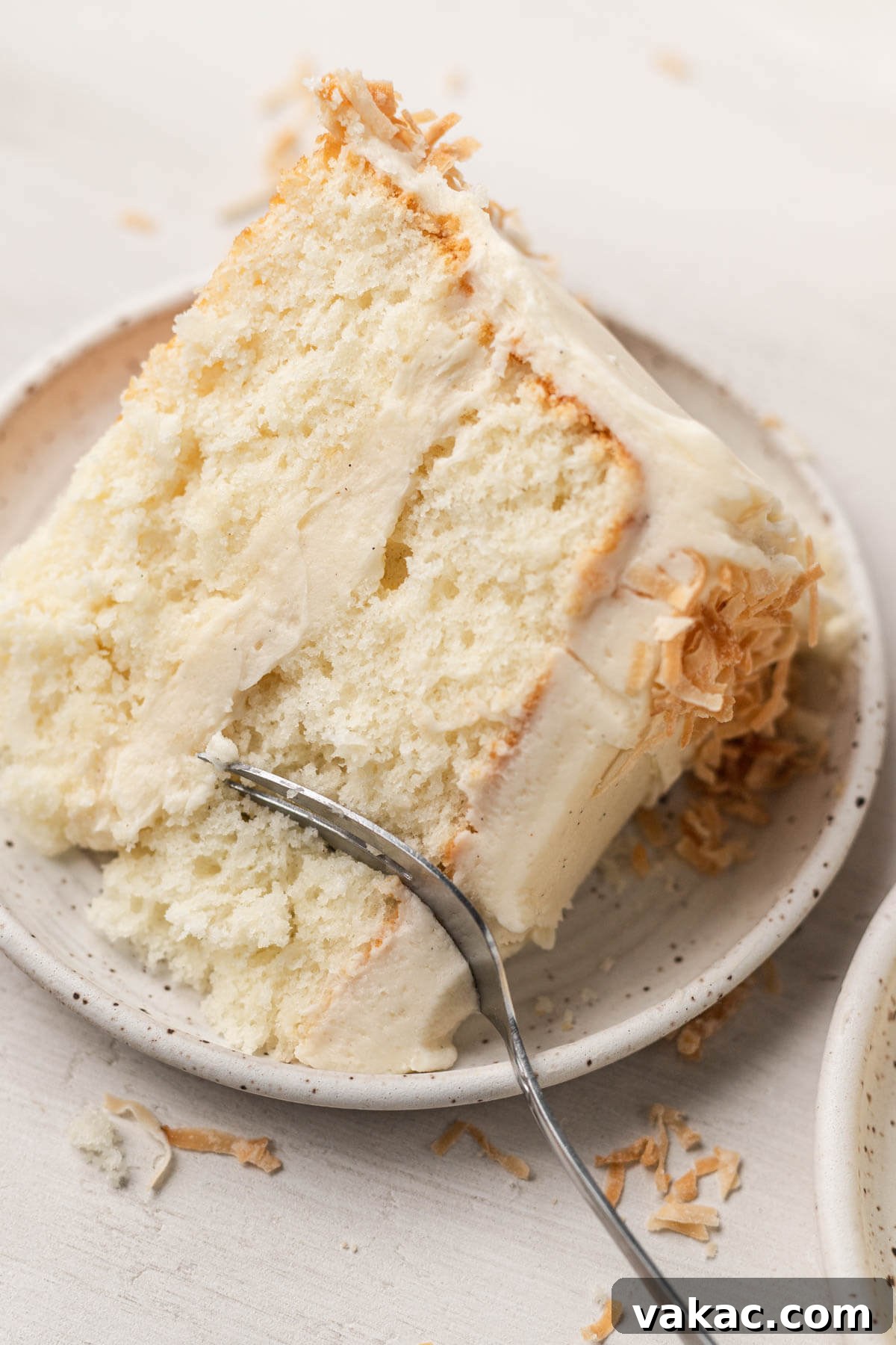 Fluffy Coconut Cloud Cake 10 A piece of frosted coconut layer cake on a white speckled dessert plate with a fork.