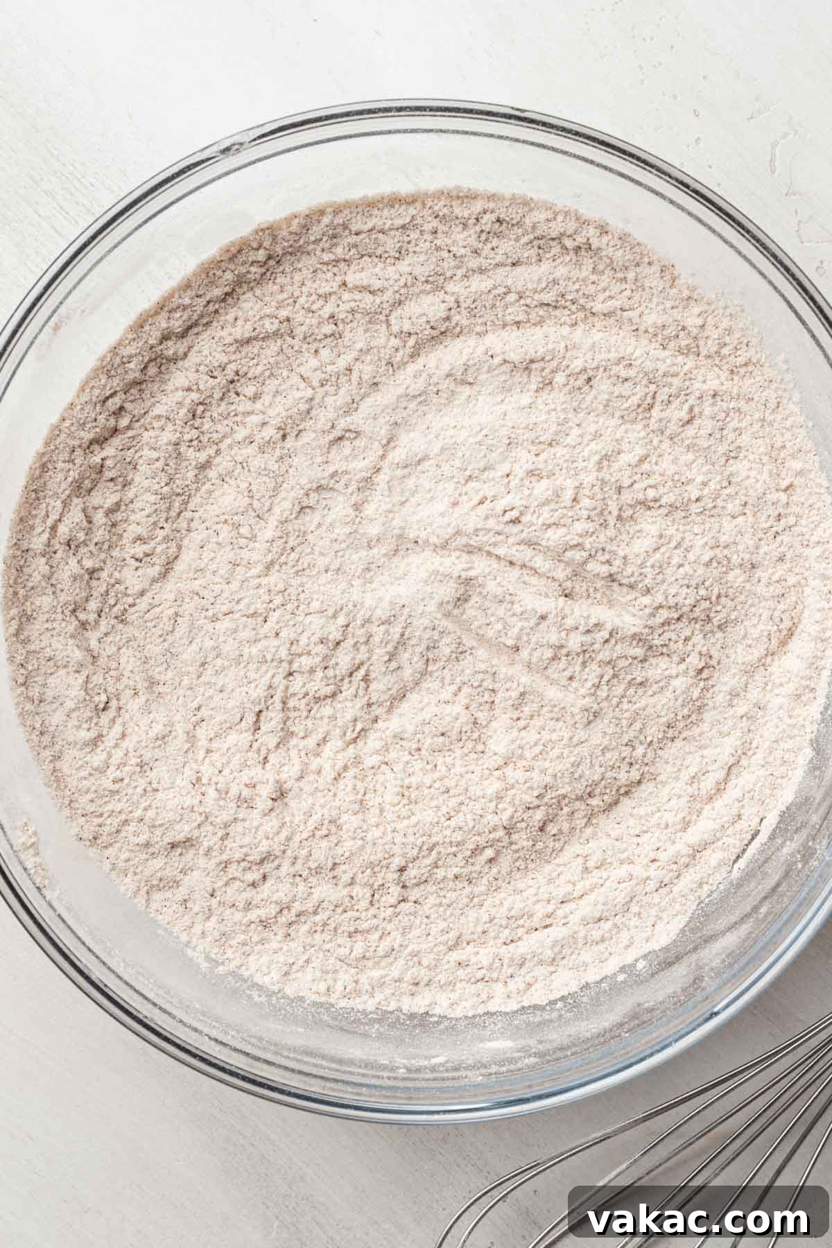 An overhead view of dry ingredients, including flour and spices, neatly arranged in a clear glass mixing bowl, ready to be combined.