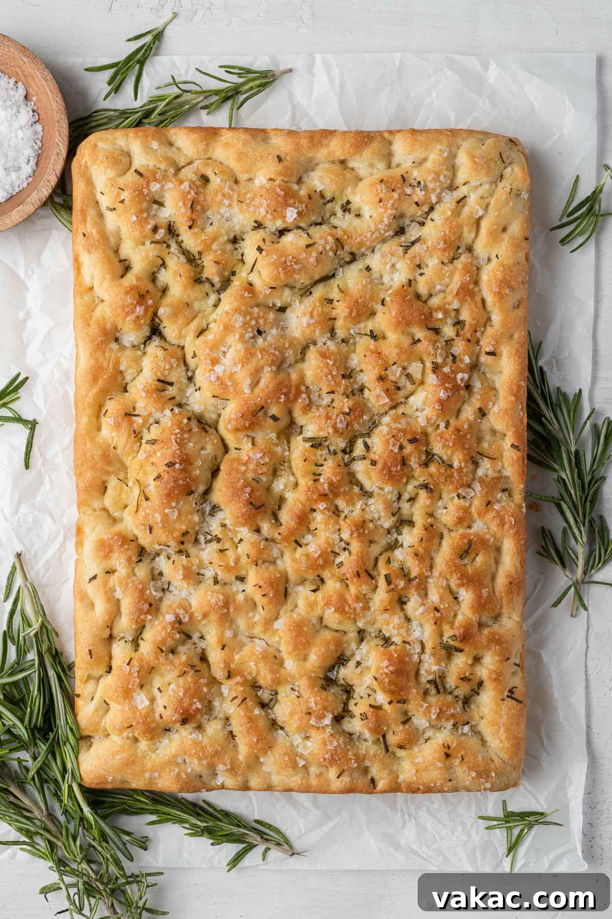 Pillowy Focaccia Perfection 6 An overhead view of a loaf of baked focaccia, surrounded by fresh rosemary sprigs and a small bowl of sea salt.
