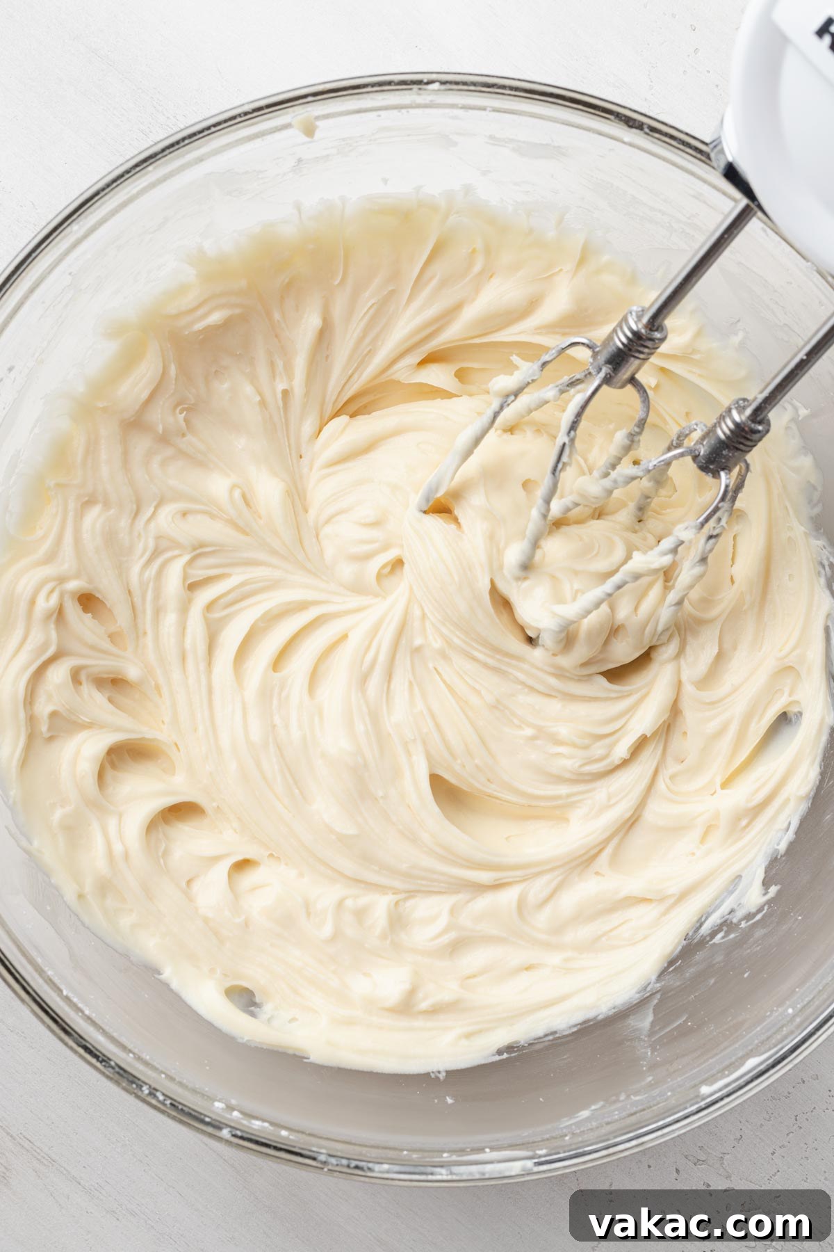 An overhead view of rich cream cheese being skillfully beaten in a clear glass mixing bowl, demonstrating a crucial step in preparing the Chantilly frosting.
