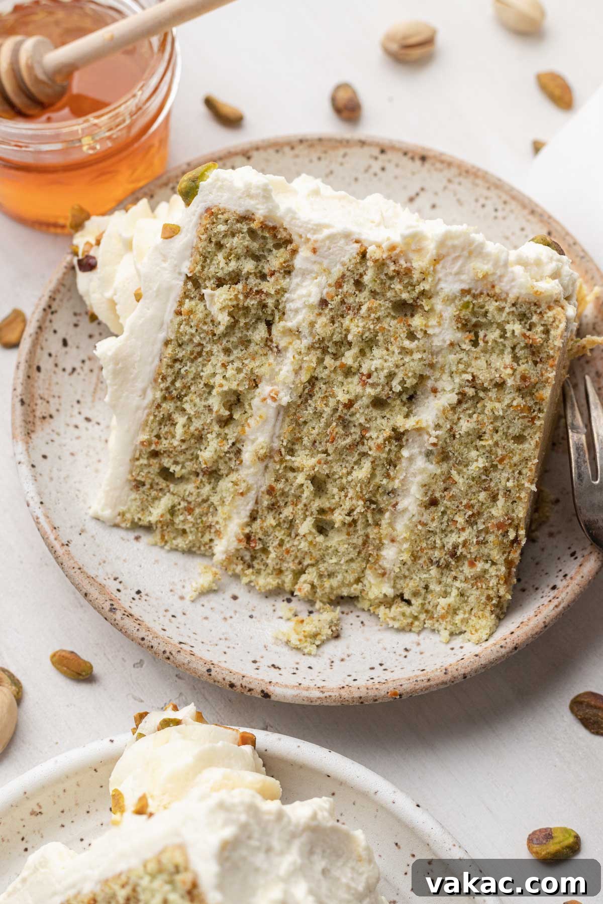 Velvet Pistachio Indulgence 5 A slice of pistachio nut cake on a speckled white dessert plate, with honey on the side.