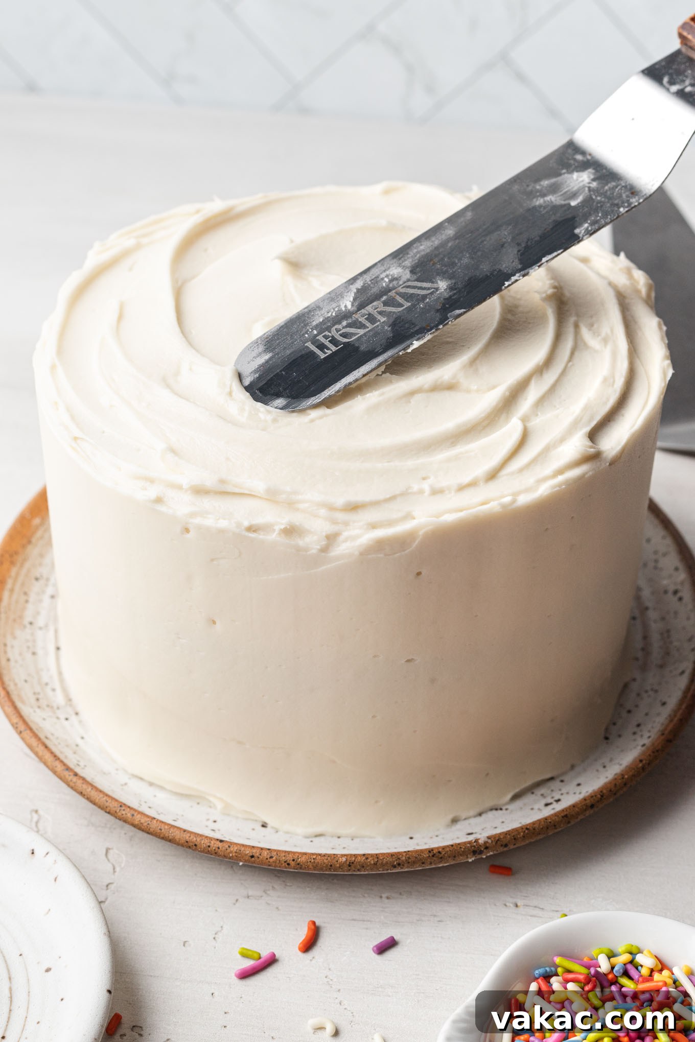 A mini vanilla cake on a speckled white plate being expertly frosted with creamy vanilla buttercream.
