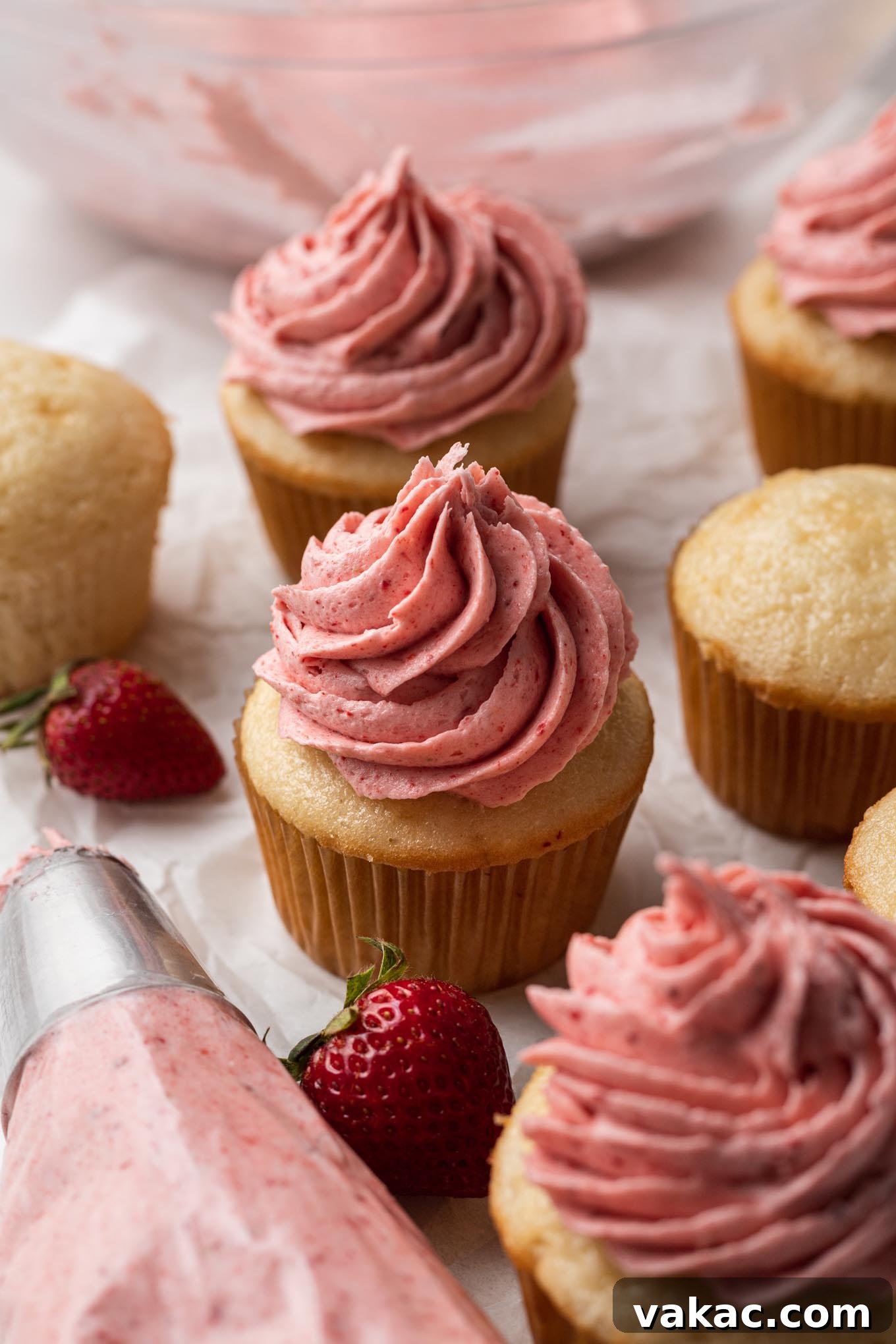 Velvet Strawberry Frosting 4 A cluster of vanilla cupcakes, some with frosting and some plain. A piping bag of frosting rests in the foreground.