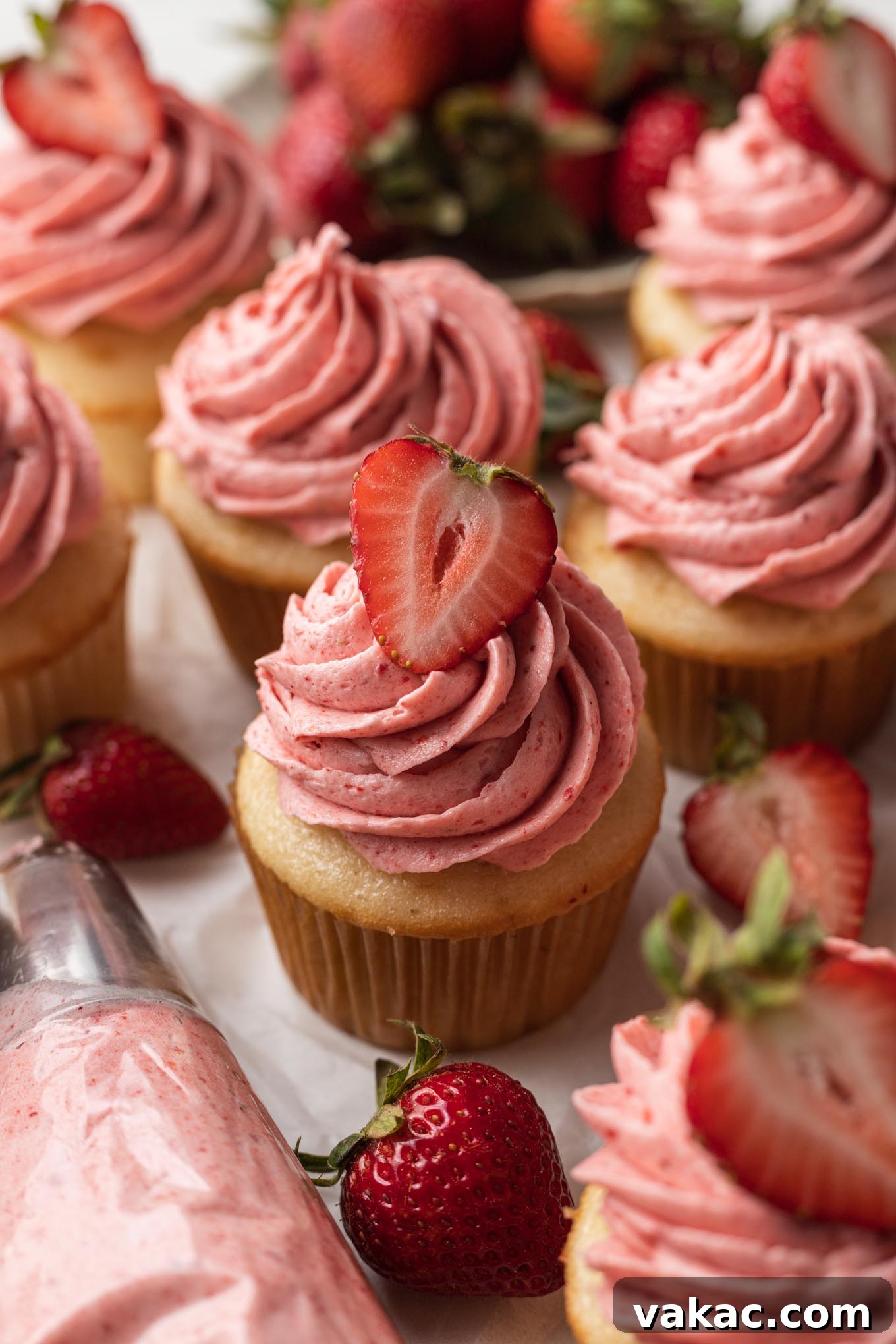 Velvet Strawberry Frosting 6 Several vanilla cupcakes topped with strawberry frosting. A piping bag with frosting rests near the cupcakes.