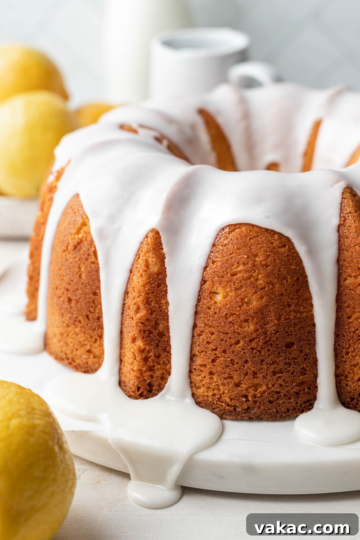 Sunshine Lemon Pound Cake 2 A glazed lemon pound cake on a white cake plate, with lemons in the foreground and background.