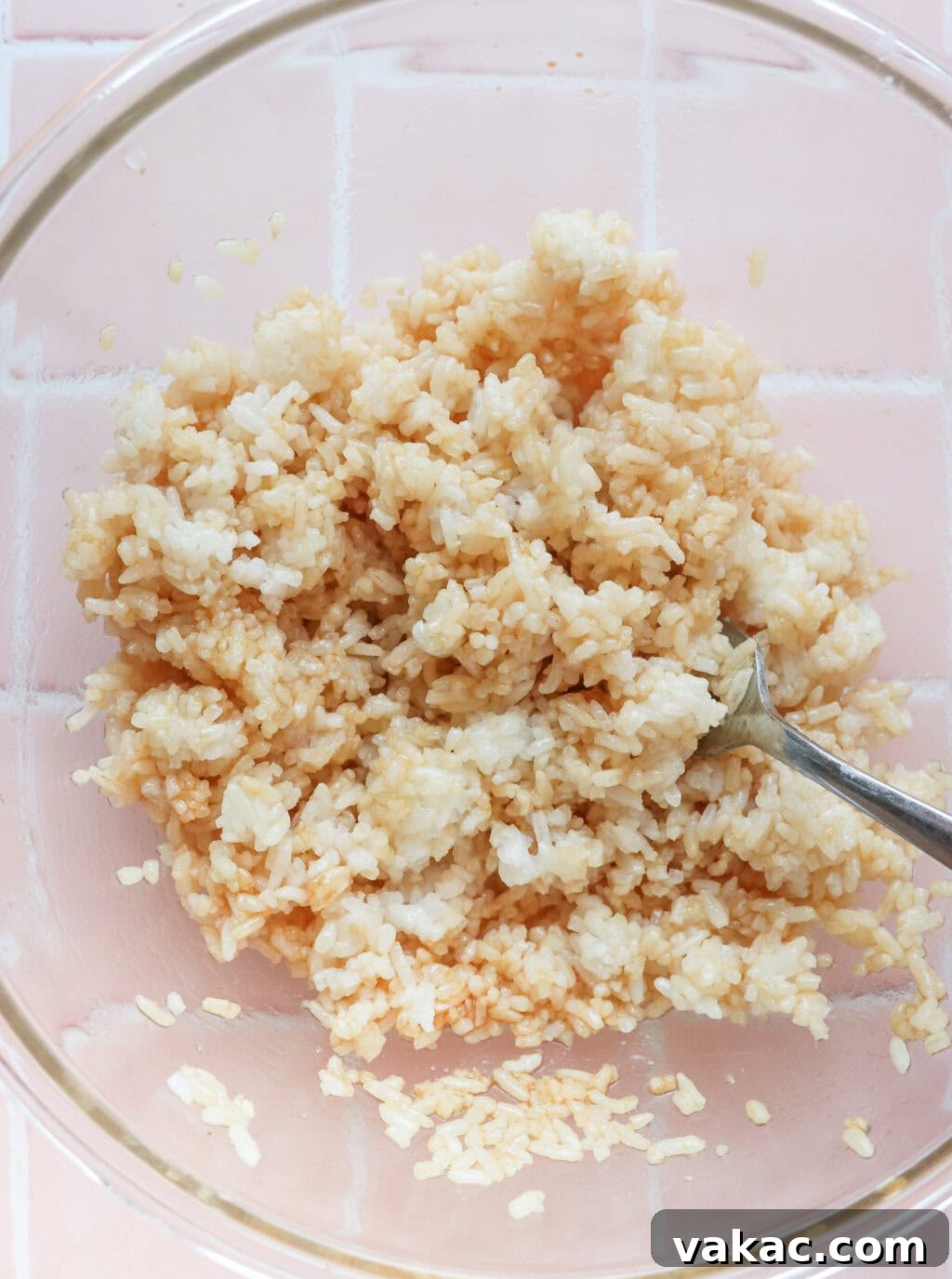 Overhead shot of cooked rice being mixed with sesame oil and soy sauce in a clear glass bowl.