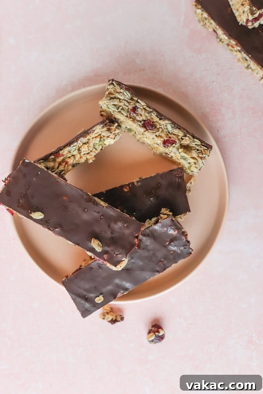 Allergy-Friendly Homemade Granola Bars 2 Five chocolate-covered granola bars with nuts and dried fruit are arranged on a round plate against a pink background. Some nuts and dried fruit pieces are scattered nearby.