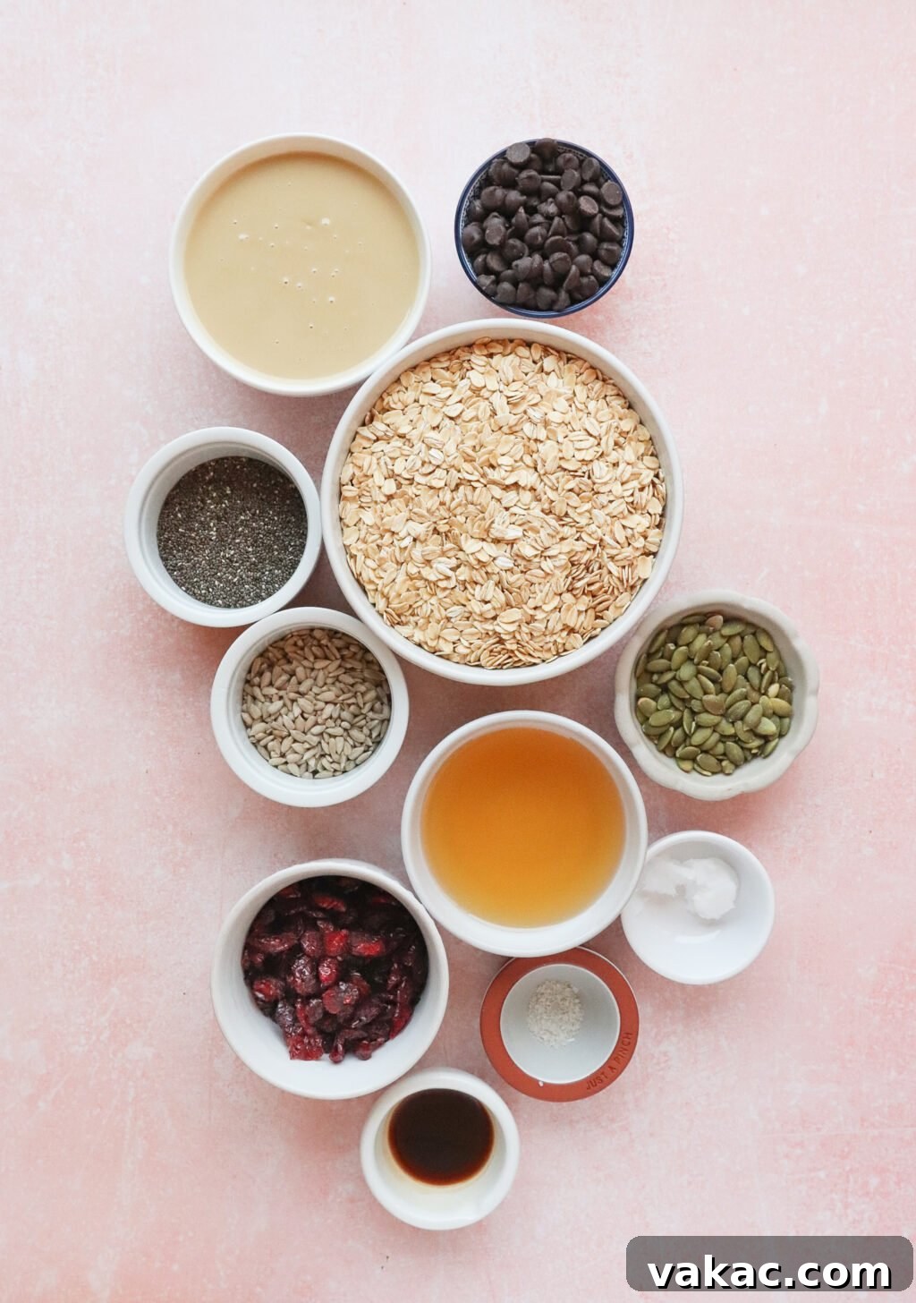 Allergy-Friendly Homemade Granola Bars 3 Various bowls on a pink surface contain ingredients: oats, chocolate chips, tahini, sunflower seeds, chia seeds, pumpkin seeds, dried cranberries, maple syrup, vanilla extract, coconut oil, and a small bowl of salt.