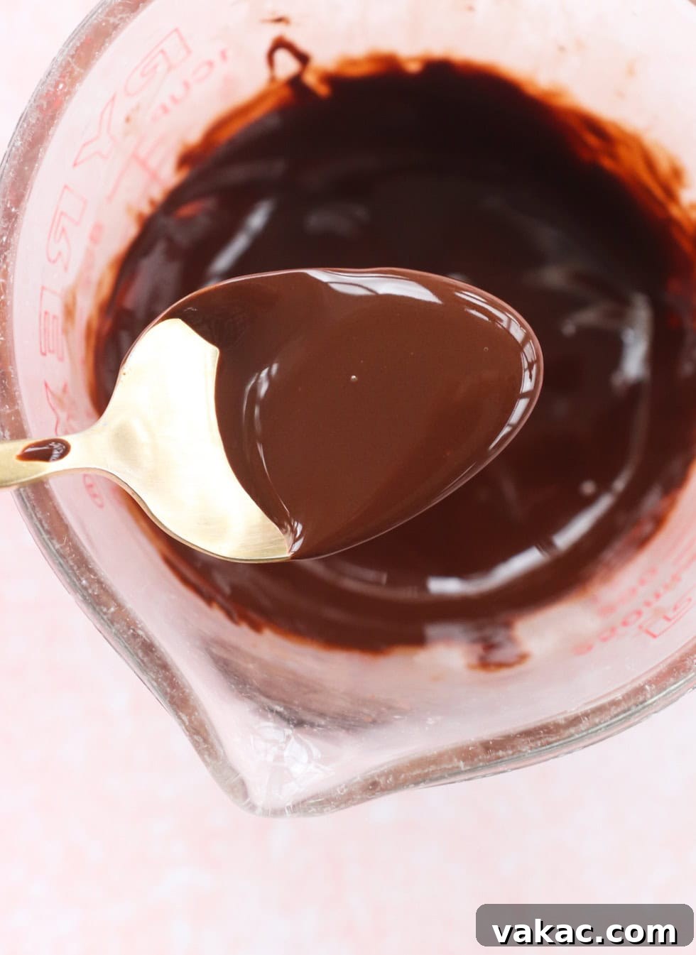 Allergy-Friendly Homemade Granola Bars 5 A spoon with melted chocolate is held above a glass measuring cup filled with more melted chocolate. The chocolate is smooth and glossy, and the measuring cup is set against a light pink background.
