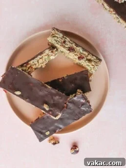 Five chocolate-covered granola bars with nuts and dried fruit are arranged on a round plate against a pink background. Some nuts and dried fruit pieces are scattered nearby.