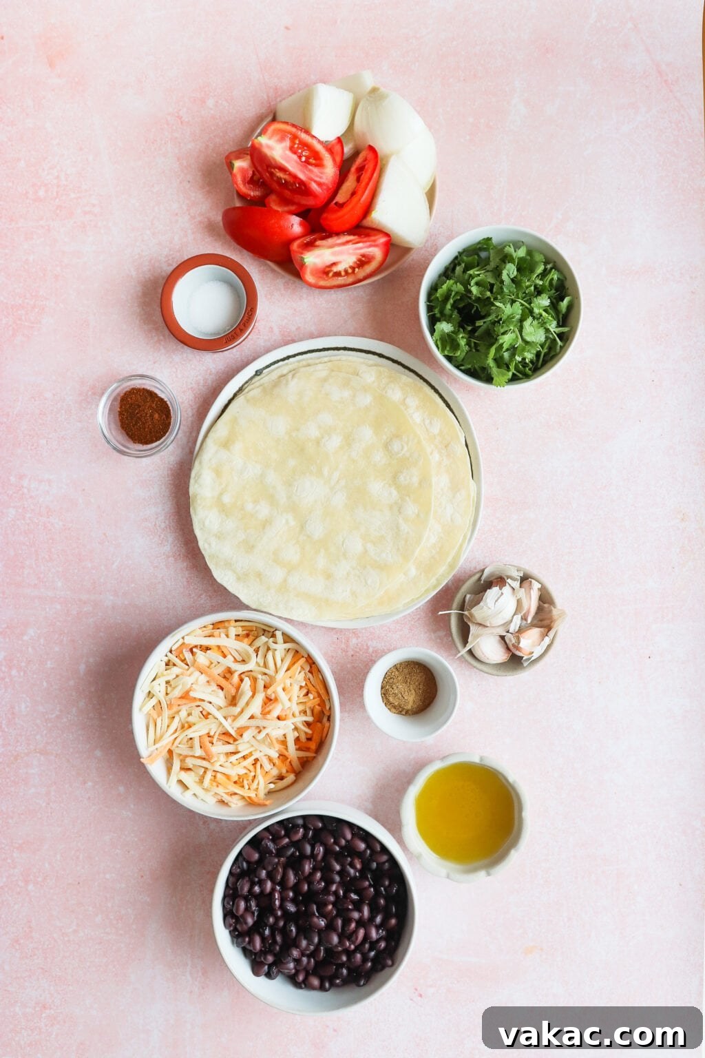 A flatlay of ingredients on a pink surface: tortillas, grated cheese, black beans, olive oil, salt, chili powder, cumin, chopped tomatoes, onions, cilantro, and garlic cloves.