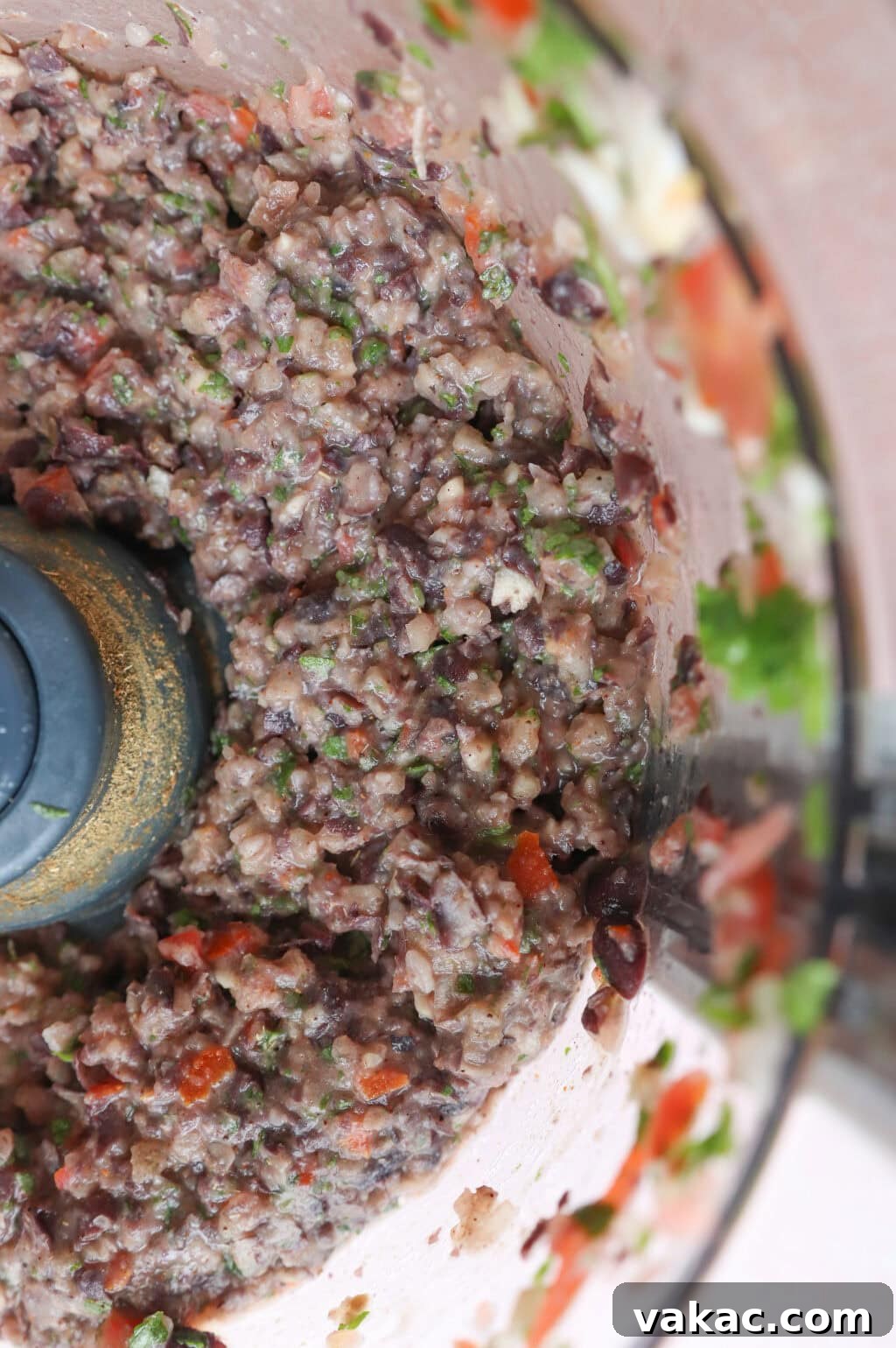 Close-up of a food processor filled with a colorful mixture of chopped ingredients, including black beans, cilantro, tomatoes, and onions, creating a chunky texture. The image shows the circular motion of the blended ingredients.