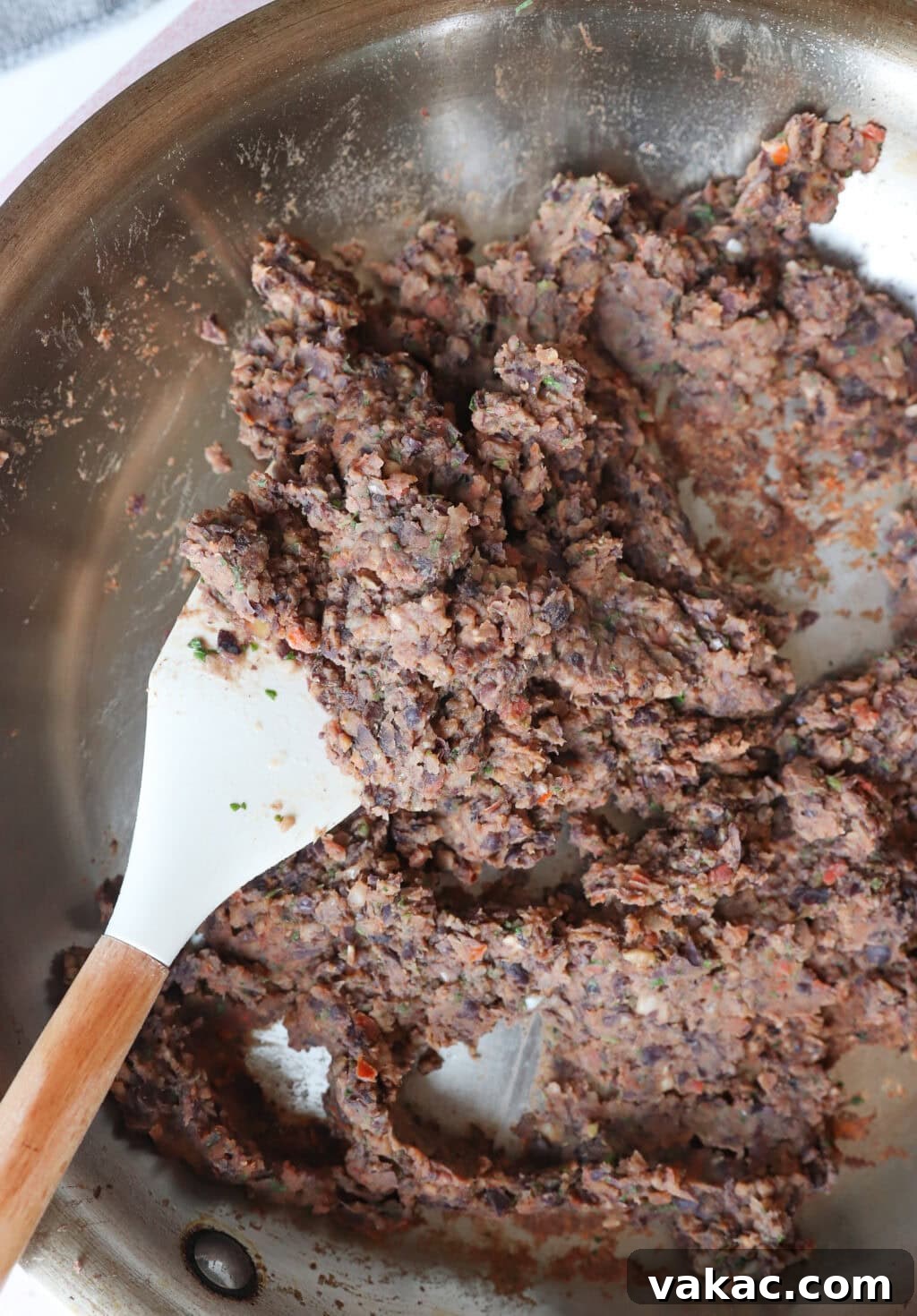 A close-up of a metal pan containing a mixture of refried black beans, being stirred with a white spatula with a wooden handle. The bean mixture appears chunky with visible herbs and spices.