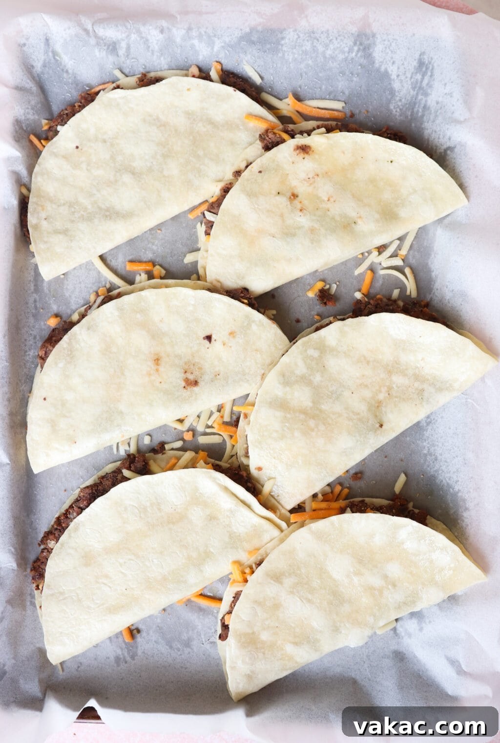 Six uncooked tacos arranged in a parchment-lined baking tray. topped with shredded cheese, ready to be baked.