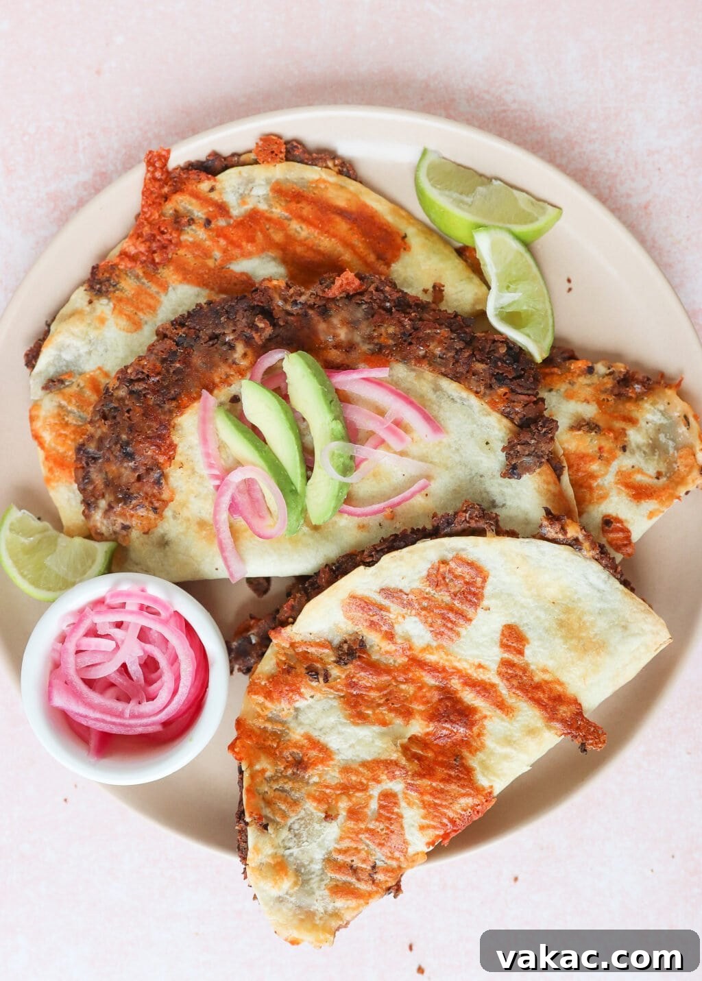 A plate of crispy quesabirria tacos with melted cheese, garnished with cucumber slices and pickled red onions. Lime wedges are on the side. A small bowl of pickled onions accompanies the dish, set against a light pink background.
