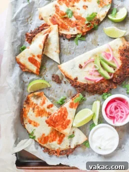 A tray of crispy tacos filled with cheese and meat, garnished with sliced avocado, pickled onions, and cilantro. Lime wedges are scattered around, with small bowls of pickled onions and sour cream on the side.