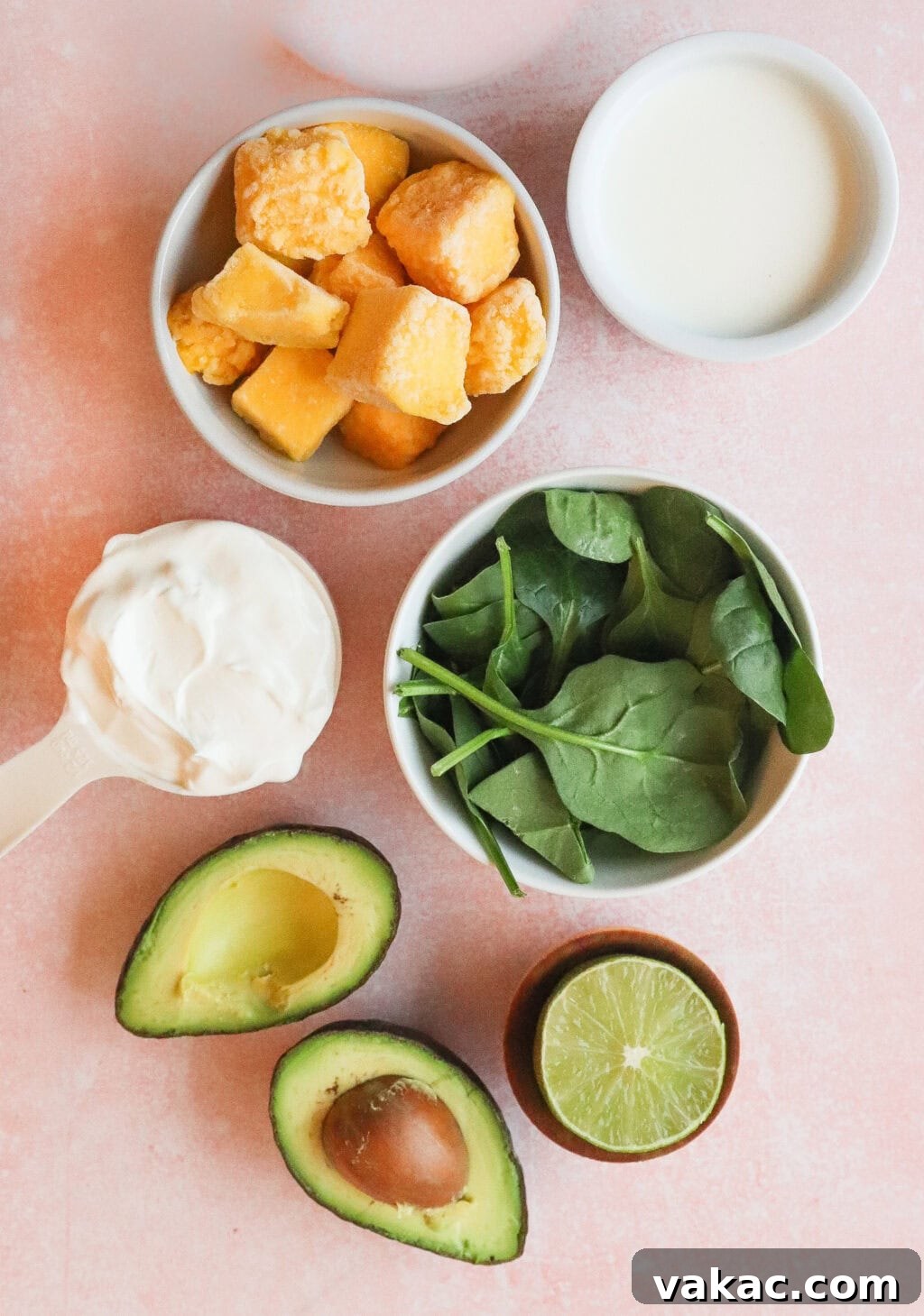 A beautifully arranged flat lay of fresh smoothie ingredients on a light pink surface, featuring a bowl of golden frozen mango chunks, a small glass of milk, a cup of creamy yogurt, a generous portion of fresh spinach leaves, a perfectly halved avocado revealing its vibrant green flesh and pit, and a halved lime, all ready for blending.