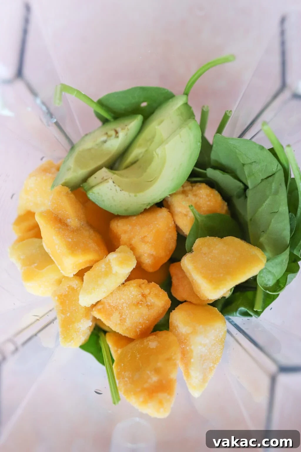 A top-down view inside a blender, showcasing the vibrant, fresh ingredients: bright green spinach leaves, creamy sliced avocado, and golden chunks of frozen mango, all layered and ready to be transformed into a delicious smoothie.