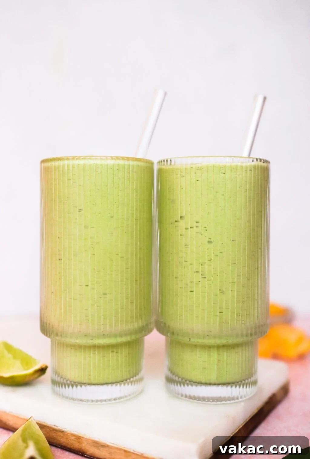 Two clear glass cups, each holding a vibrant green smoothie and a matching clear straw, are placed side by side on a clean white surface. Fresh lime wedges and bright orange slices are artfully arranged nearby, with a softly blurred background providing a gentle contrast to the crisp foreground elements, inviting a refreshing sip.