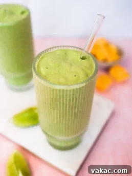 A glass of green smoothie with a clear straw sits on a white board, with lime wedges and a bowl of orange fruit pieces in the background. Another glass of green smoothie is partially visible beside it.