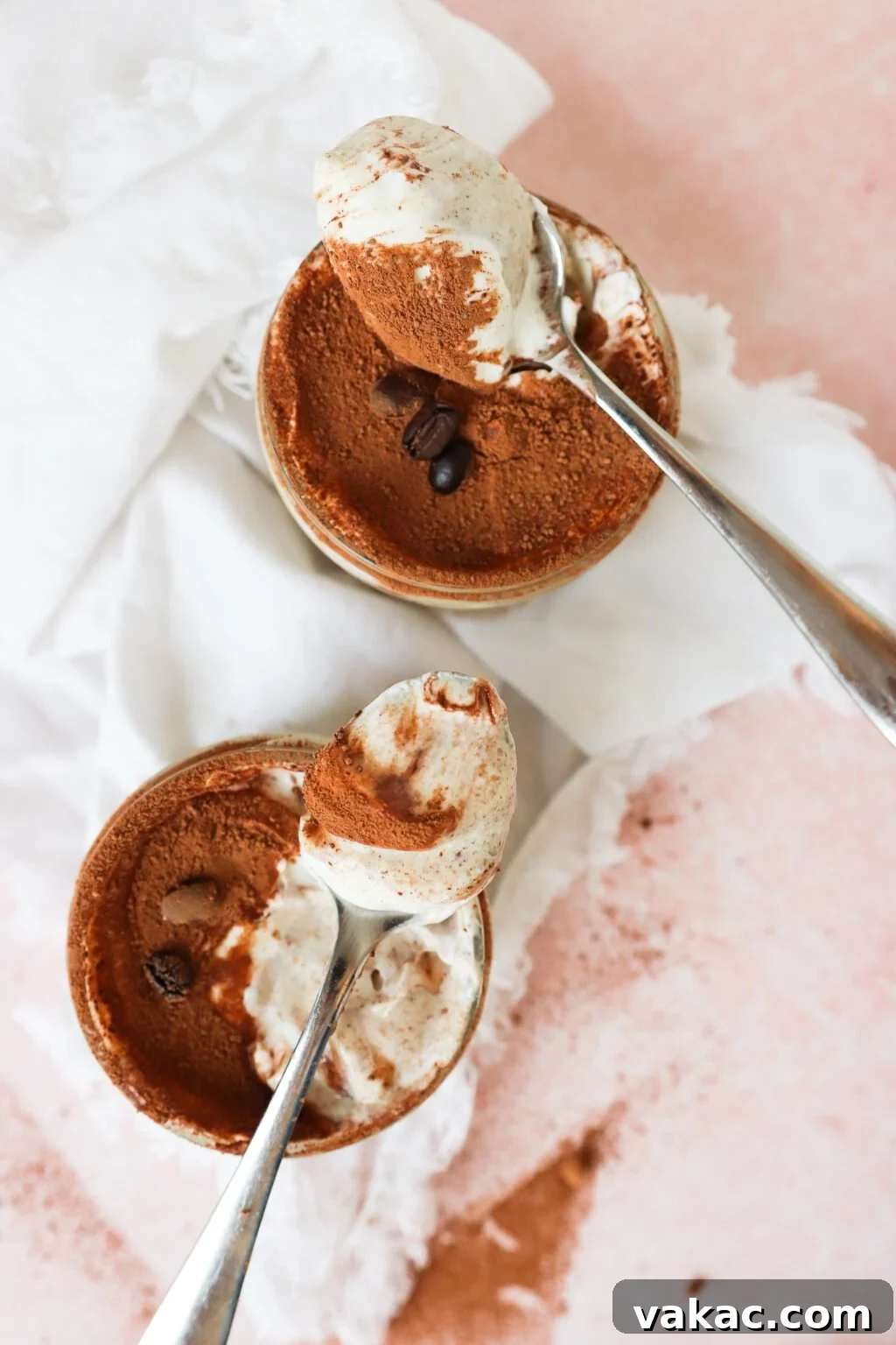 Dreamy Protein Tiramisu Chia Pudding with Yogurt 2 Two metal spoons holding creamy tiramisu are placed above two small ramekins filled with tiramisu topped with cocoa powder and coffee beans, set on a white cloth and a pink surface.