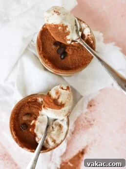 Two metal spoons holding creamy tiramisu are placed above two small ramekins filled with tiramisu topped with cocoa powder and coffee beans, set on a white cloth and a pink surface.
