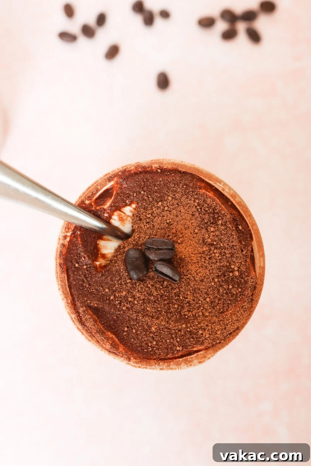 Dreamy Protein Tiramisu Chia Pudding with Yogurt 4 A top view of a chocolate dessert or drink topped with cocoa powder and coffee beans, with a spoon inside and scattered coffee beans in the background on a light pink surface.