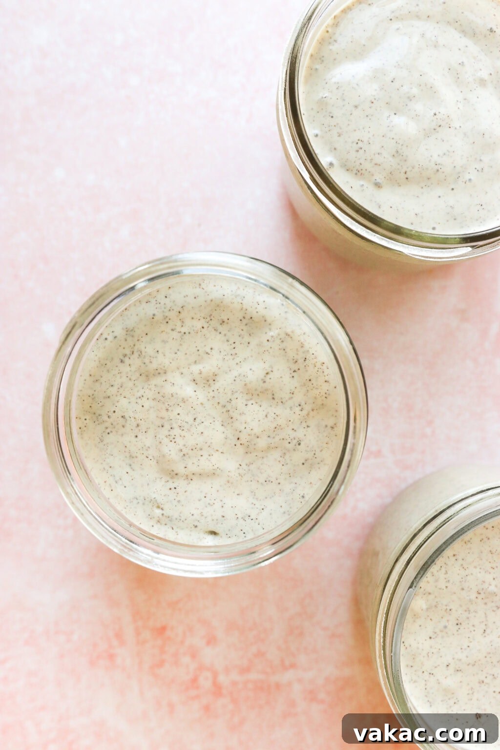 Dreamy Protein Tiramisu Chia Pudding with Yogurt 7 Three glass jars filled with a creamy, pale mixture speckled with tiny black vanilla bean flecks are arranged on a soft pink surface, viewed from above.
