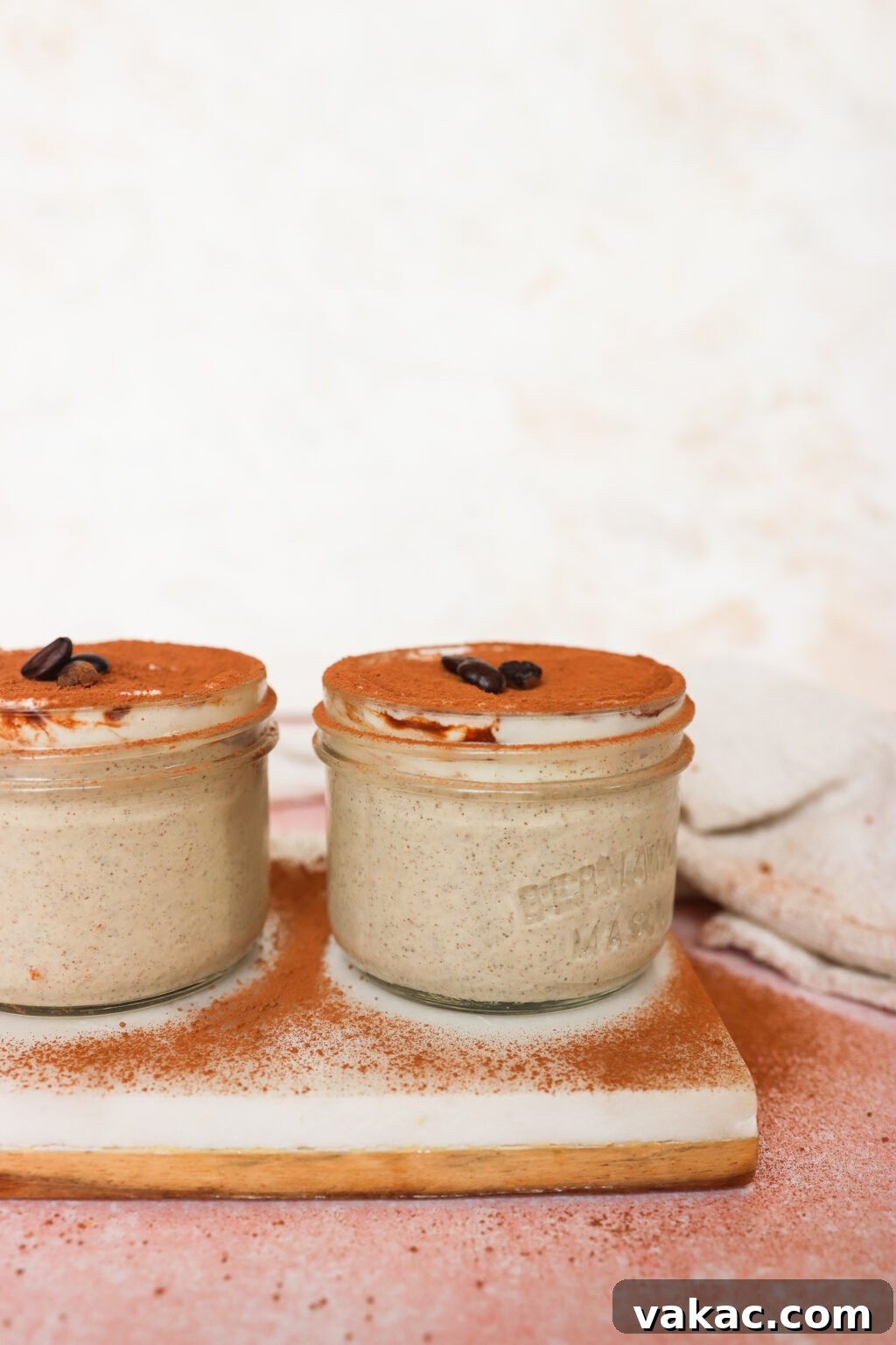 Dreamy Protein Tiramisu Chia Pudding with Yogurt 9 Two small glass jars filled with creamy tiramisu, topped with cocoa powder and a coffee bean, sit on a white wooden board. The background is softly blurred in beige and pink tones.
