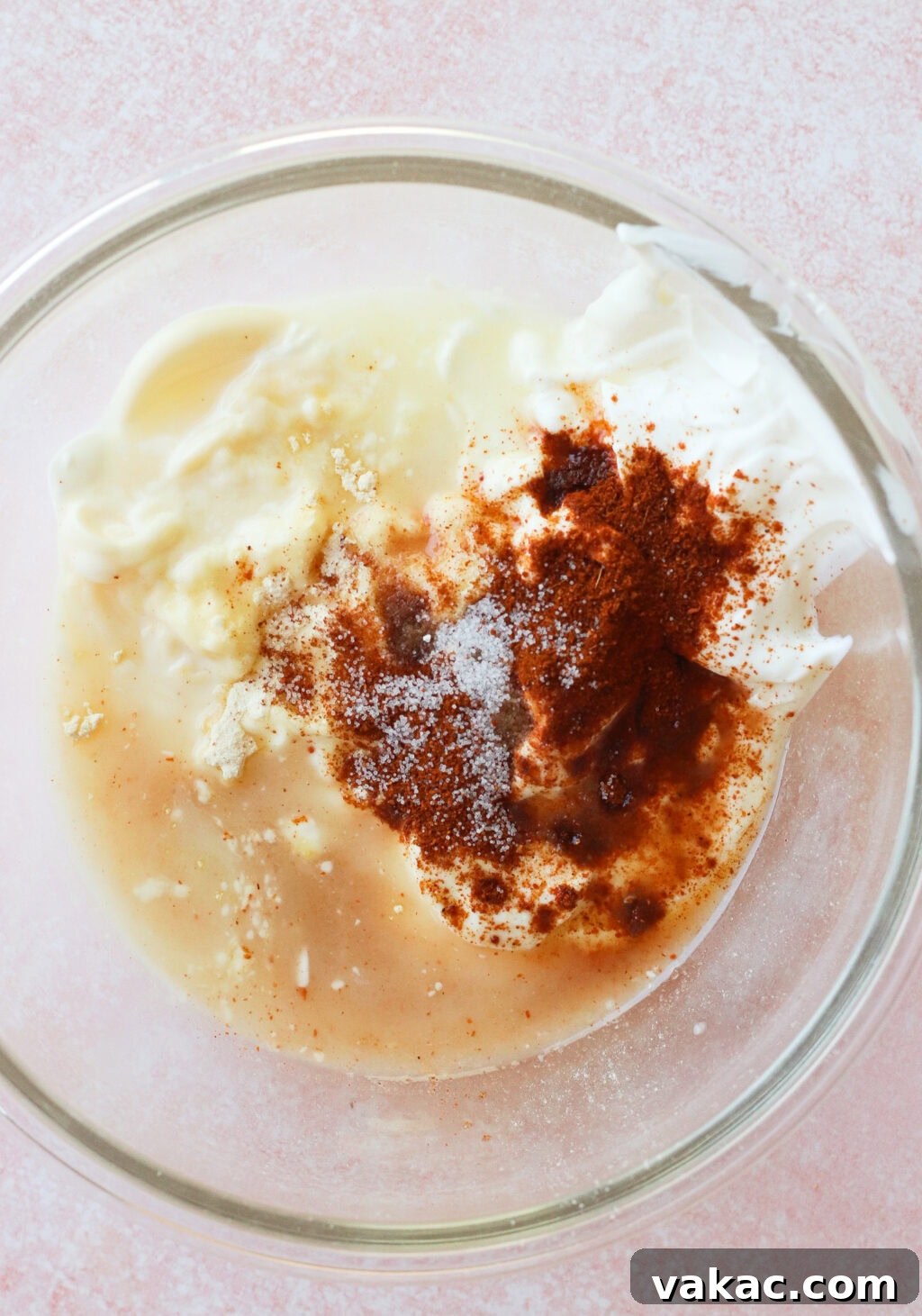A glass bowl containing sour cream, mayonnaise, spices, and minced garlic, all unmixed, on a light pink textured surface.