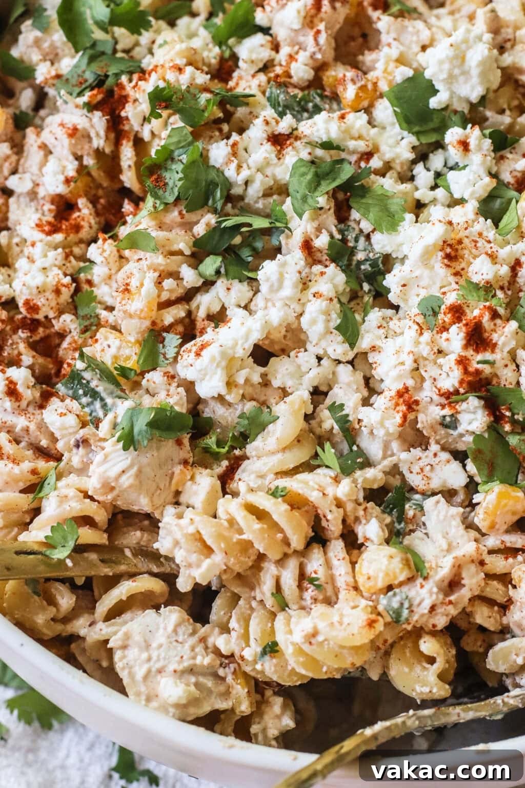 A close-up of a pasta salad made with rotini, shredded chicken, crumbled cheese, chopped cilantro, and sprinkled with paprika. The dish appears creamy and is served in a white bowl.
