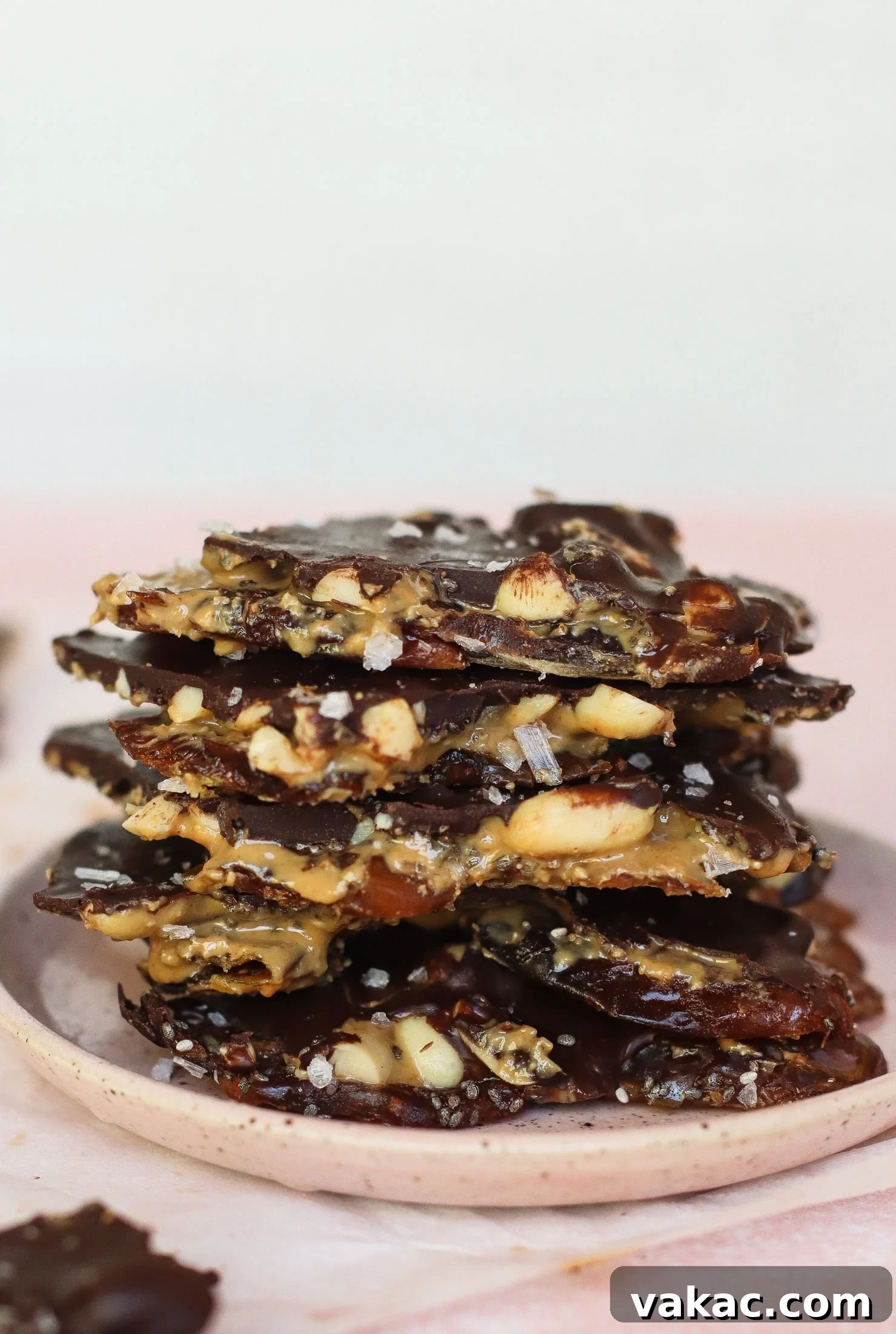 Snickers Inspired Peanut Butter Date Bark 2 A stack of chocolate-covered nut brittle pieces on a pink plate, sprinkled with flaky sea salt. The brittle shows layers of nuts and dates, with a glossy chocolate coating.