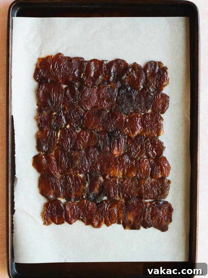 Snickers Inspired Peanut Butter Date Bark 5 A rectangular layer of homemade fruit leather on parchment paper, placed on a dark baking tray. The fruit leather appears shiny, dark brown, and slightly wrinkled.
