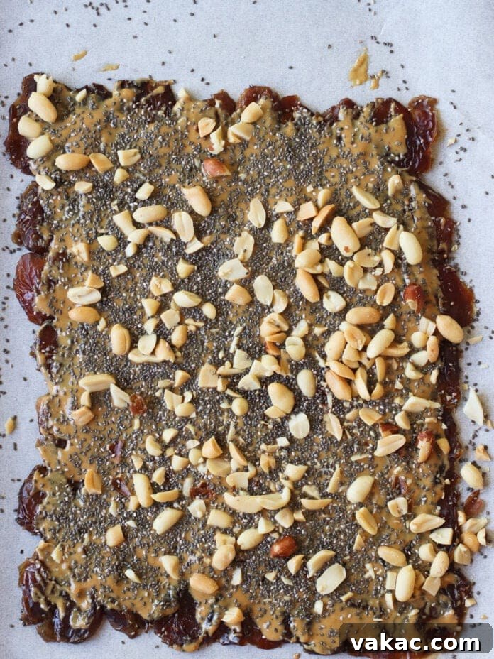 Snickers Inspired Peanut Butter Date Bark 6 A rectangular sheet of parchment paper topped with a layer of dates, spread with peanut butter, and sprinkled with chia seeds and chopped peanuts.