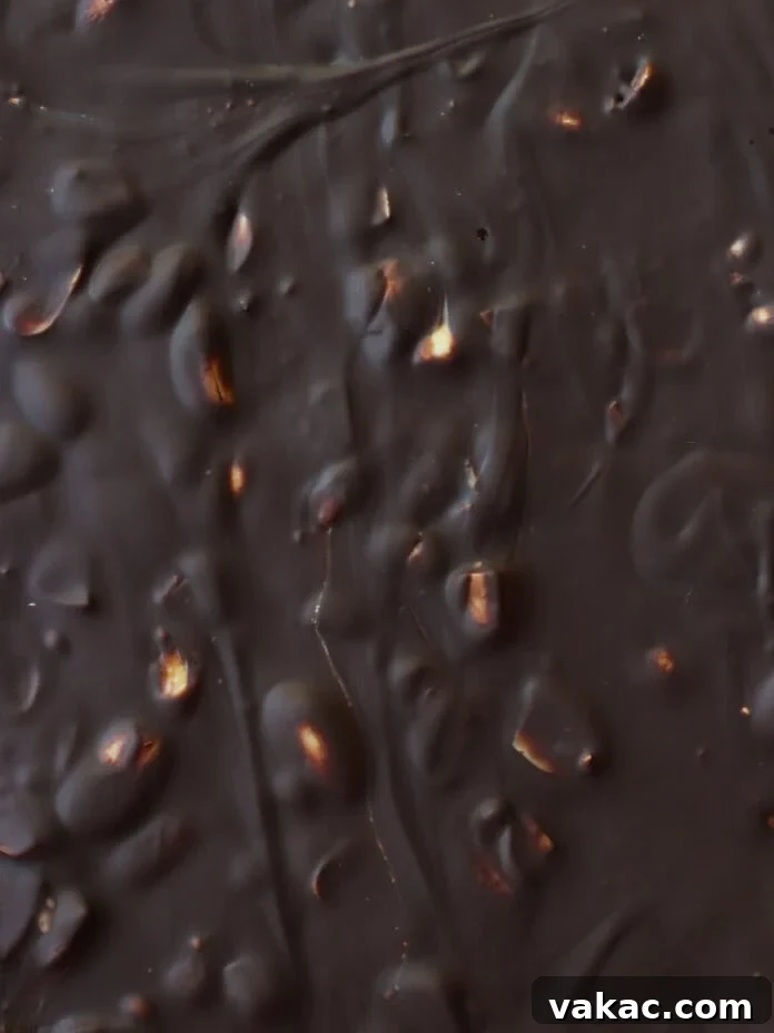 Snickers Inspired Peanut Butter Date Bark 8 Close-up of a dark chocolate bar with visible nuts or crisped rice pieces embedded, creating a bumpy and textured surface with swirls and glossy areas.